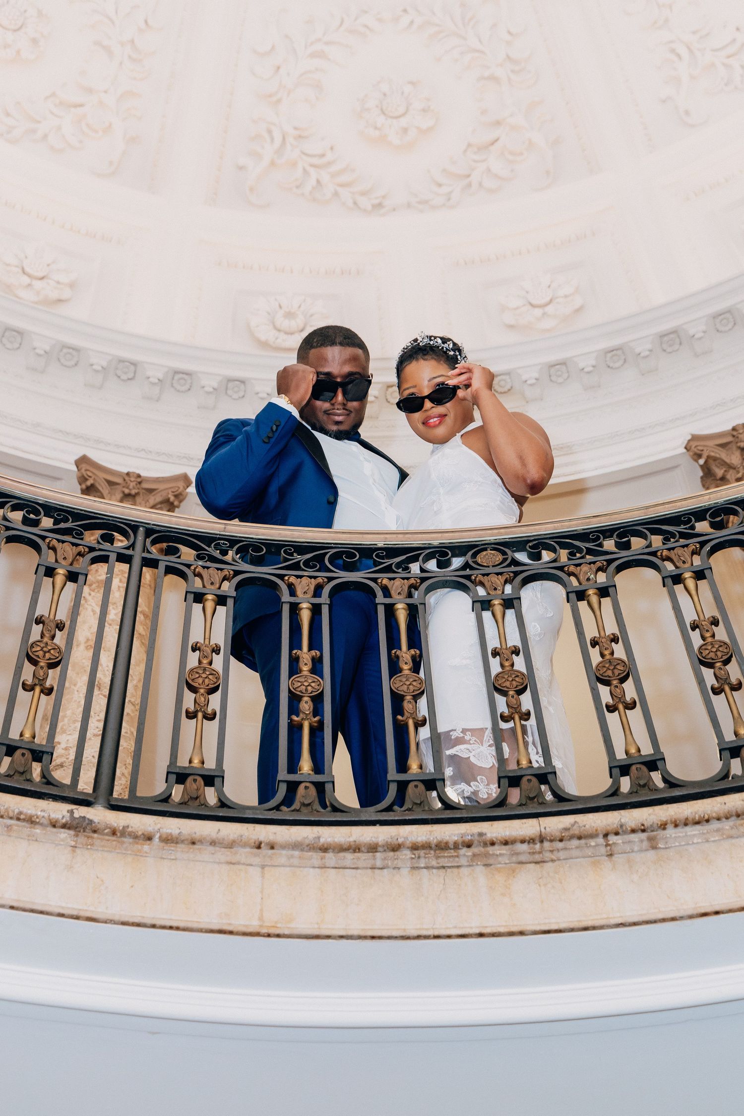 Stylish bride and groom pose with sunglasses on decorative balcony in grand wedding venue.
