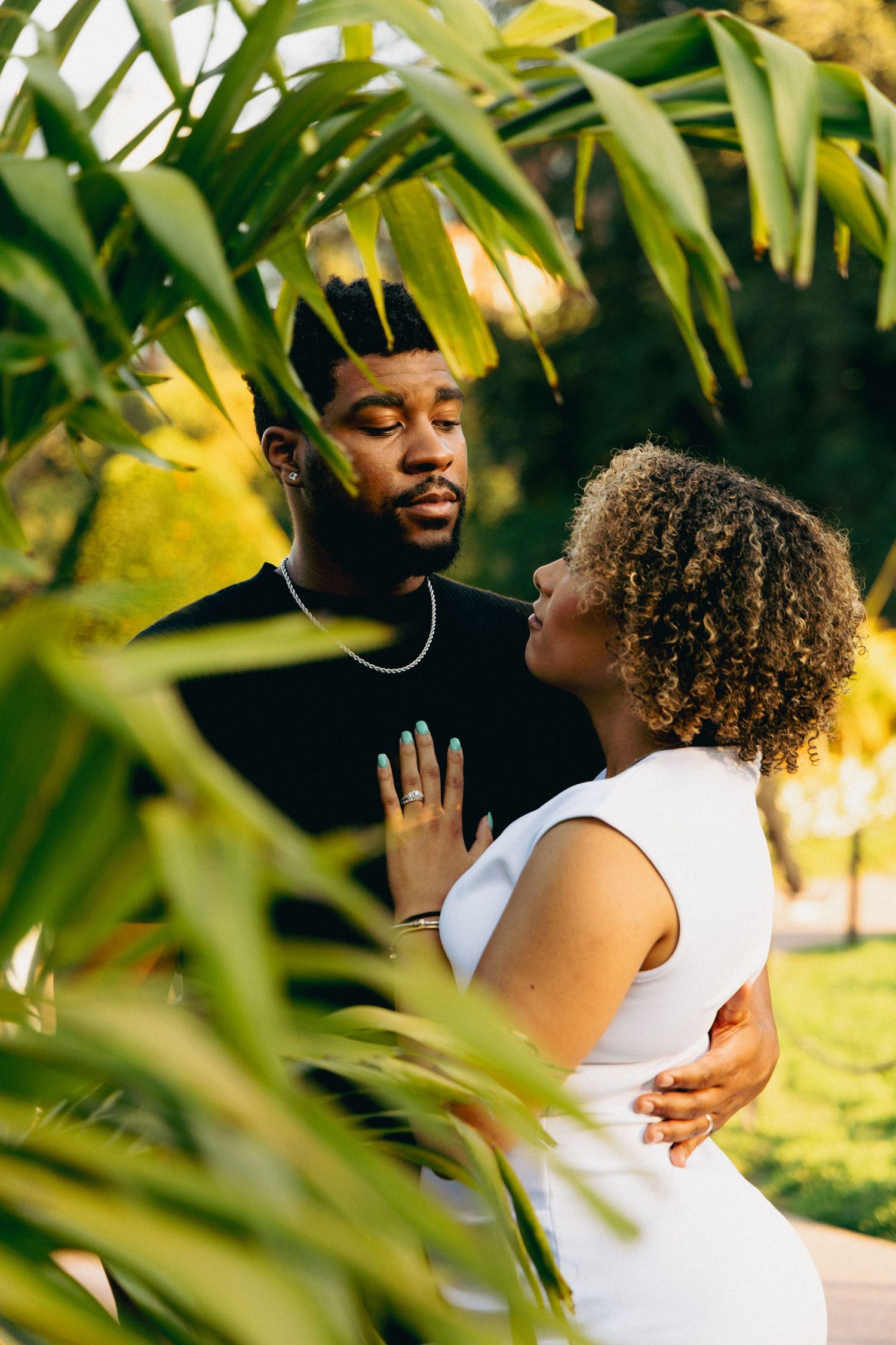 A couple shares a tender moment framed by tropical green leaves in a sunlit park setting.