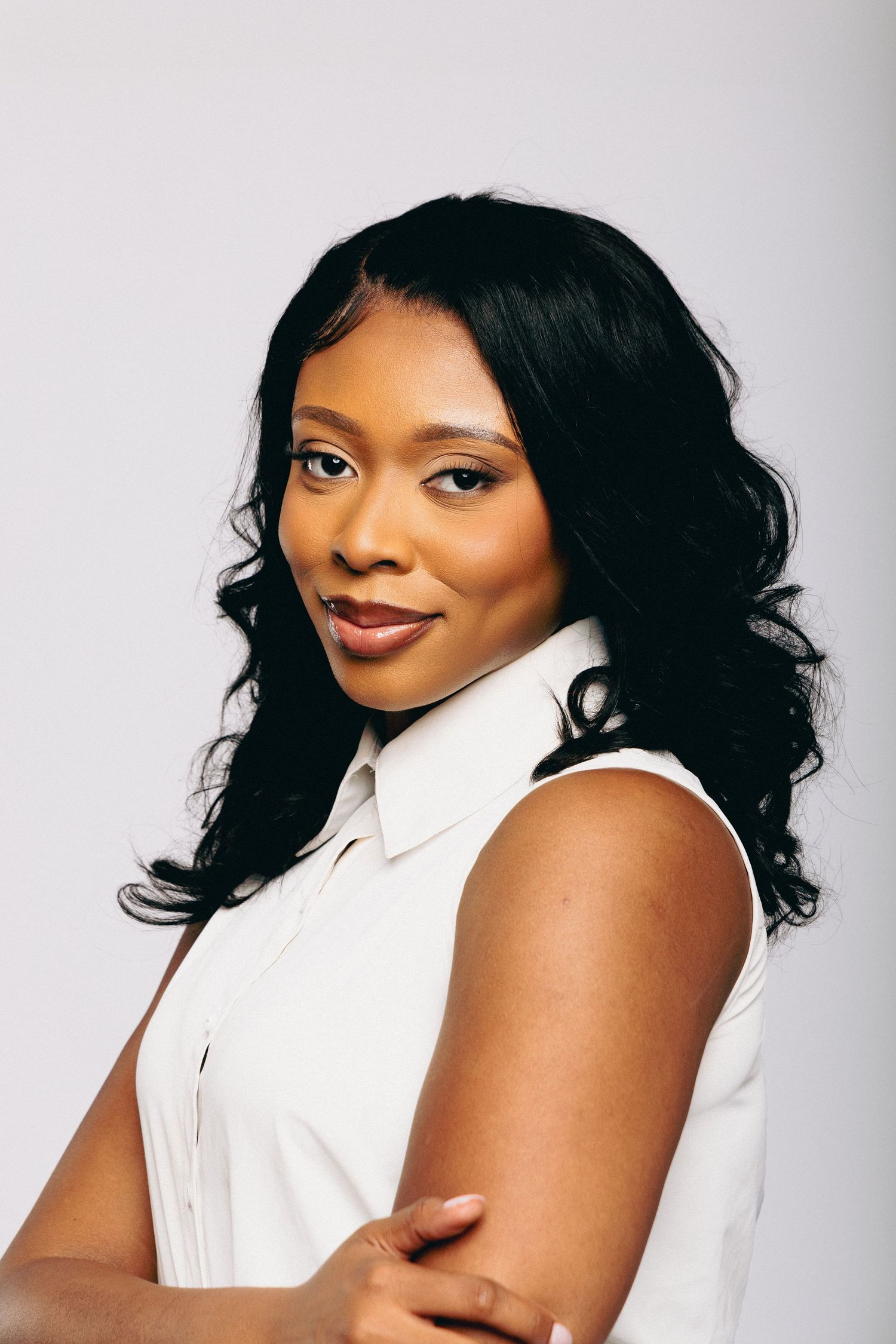 A young Black woman with curly black hair smiles confidently in a white sleeveless shirt against a gray background.