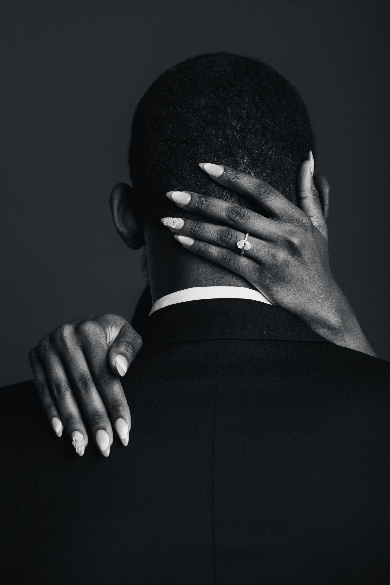 Black and white photo of a woman's hands with engagement ring embracing a man in a suit from behind.