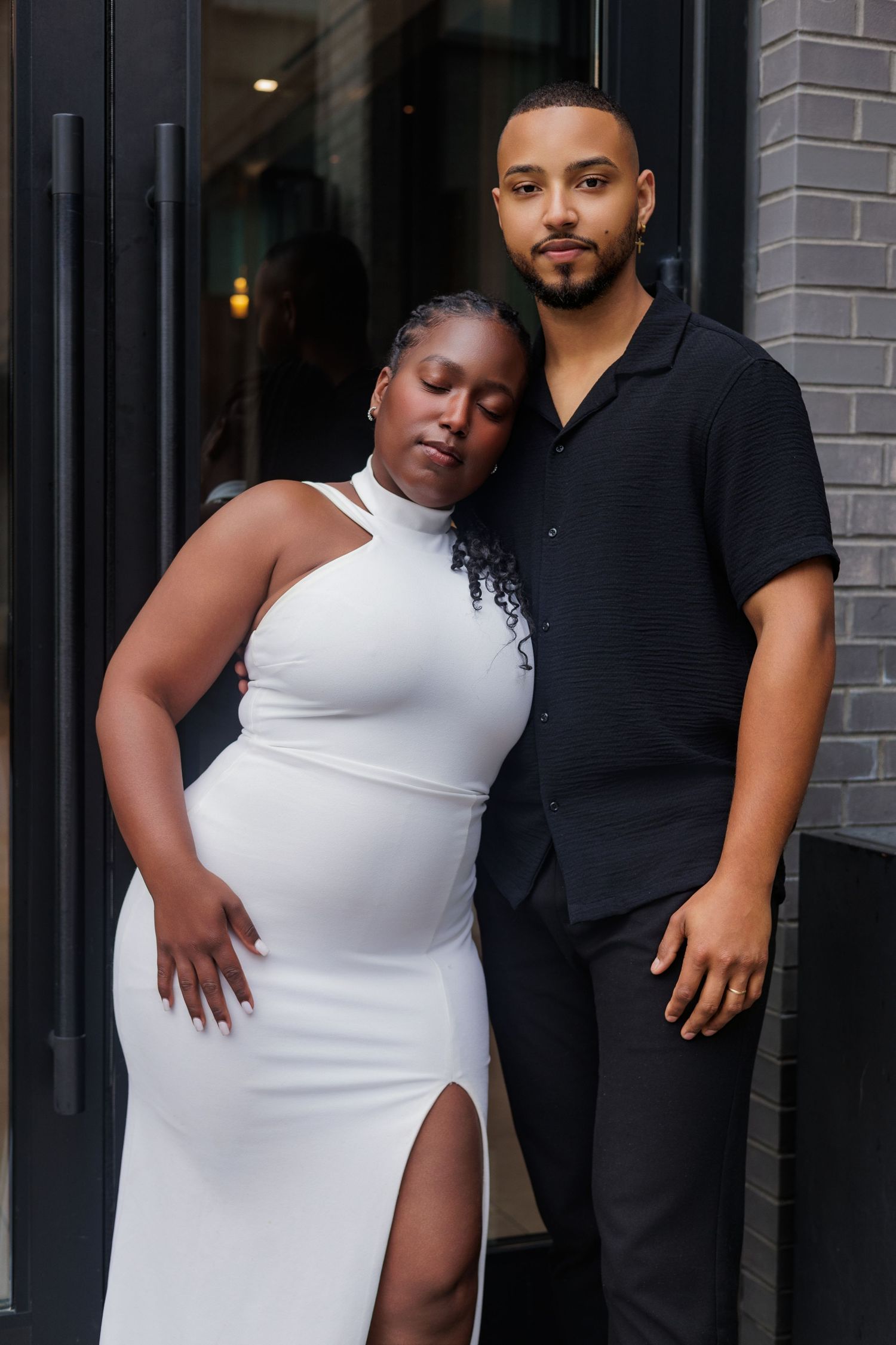A pregnant Black woman in a white dress leans on her partner in a black shirt during a maternity photo shoot.