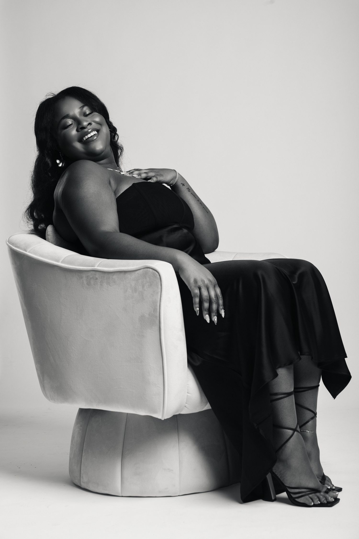 Black and white portrait of a smiling woman in elegant black dress seated in a velvet chair, studio photography.
