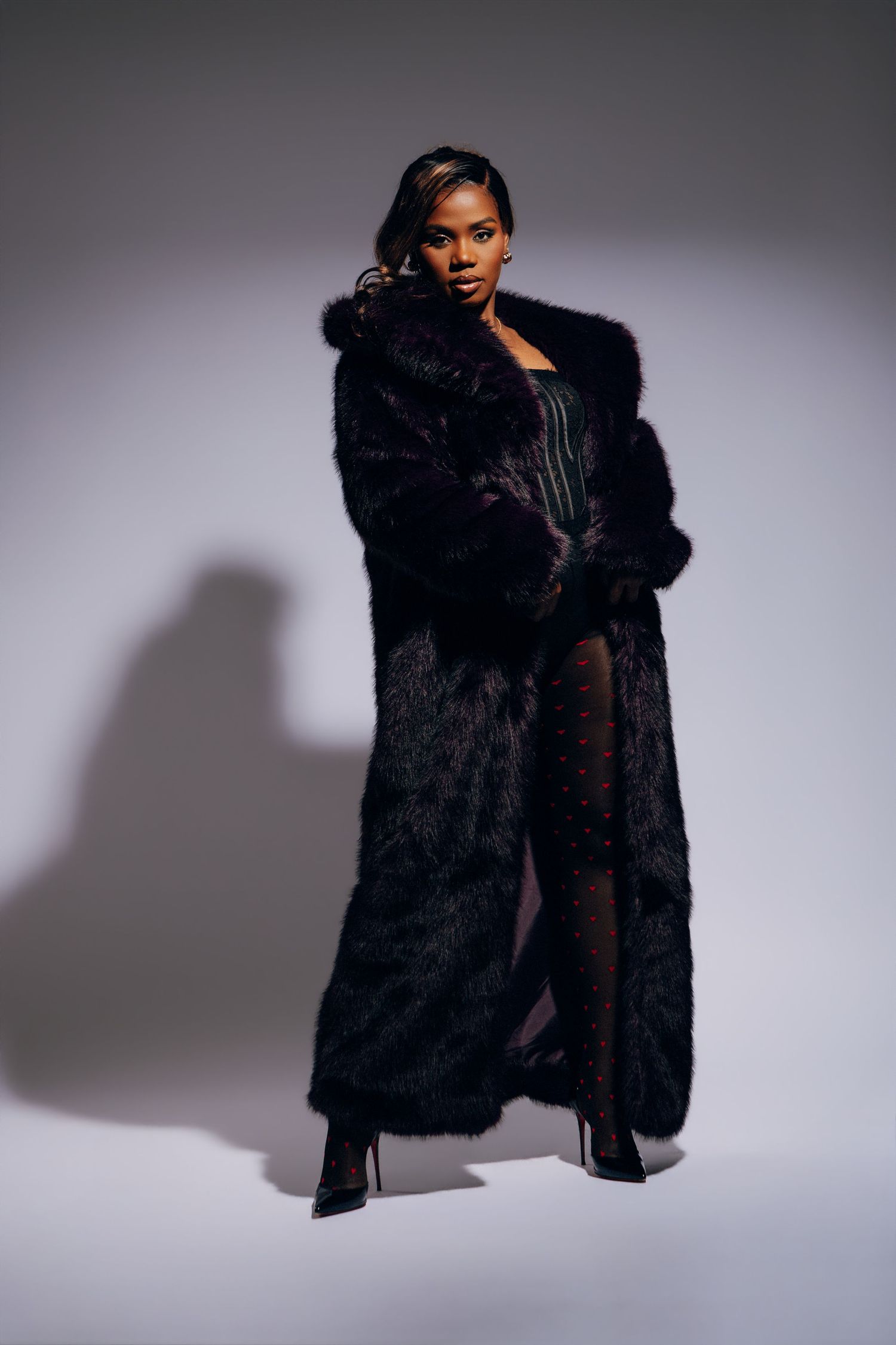 Stylish Black woman modeling a full-length black faux fur coat with black heels on grey background.