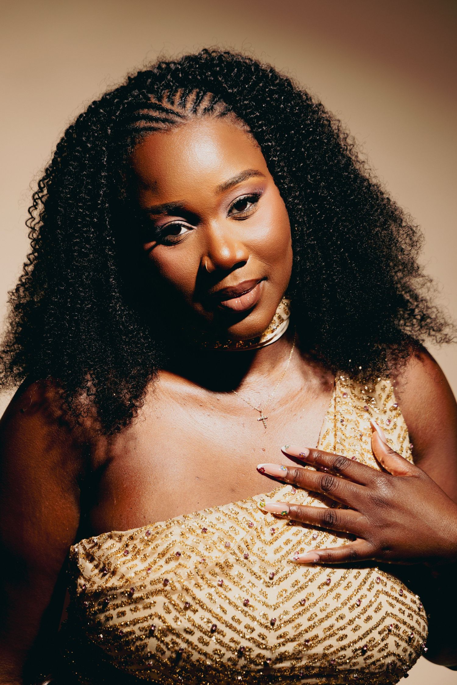 A young Black woman with curly braided hair wearing a gold sequin dress and cross necklace poses confidently.