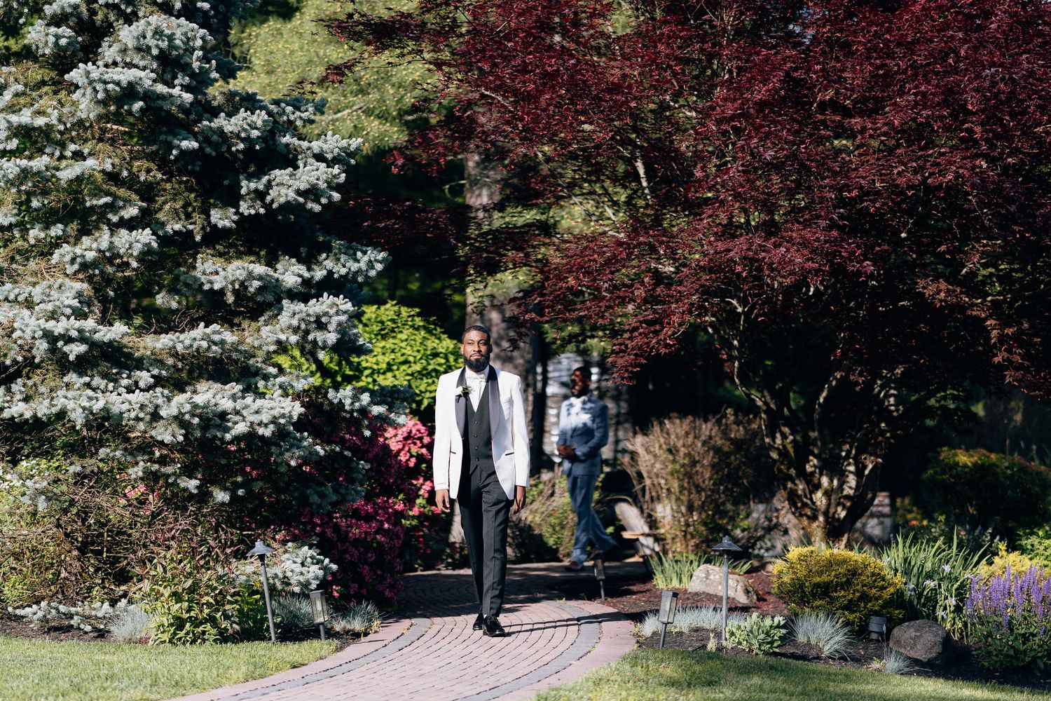 Two people walking along a brick path through a lush garden with colorful trees and flowers on a sunny day.