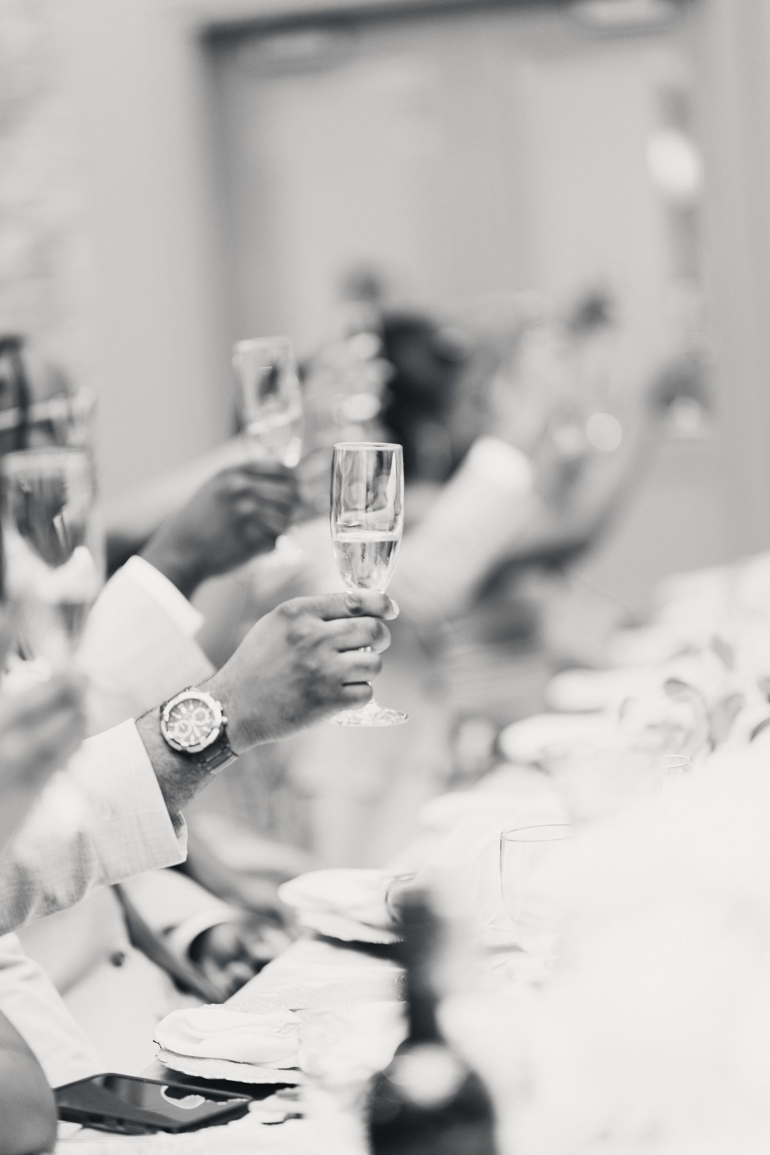 Black and white photo of wedding guests raising champagne flutes in a celebratory toast at reception.