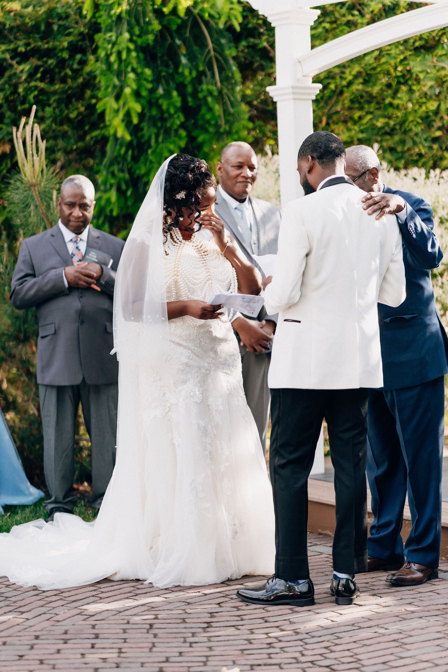 Bride and groom exchanging vows at an outdoor wedding ceremony with officiant and guests under a white arbor.