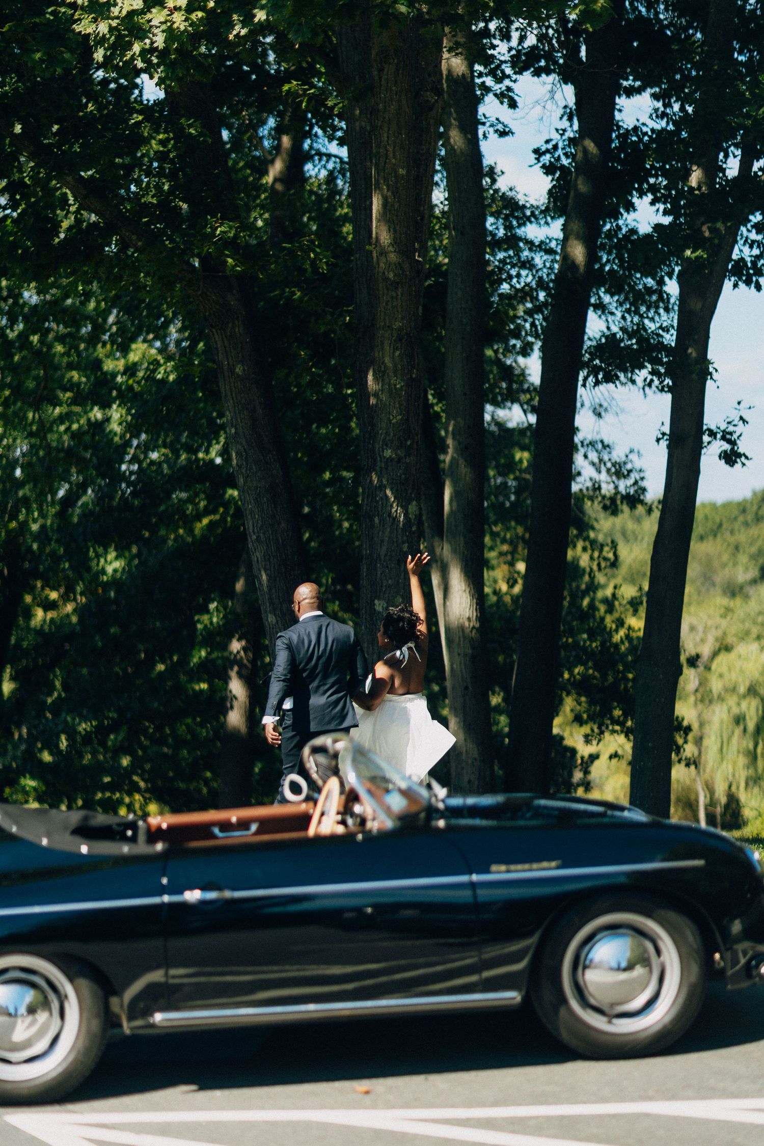 Bride and groom strolling past a classic black vintage convertible car lined with tall trees.