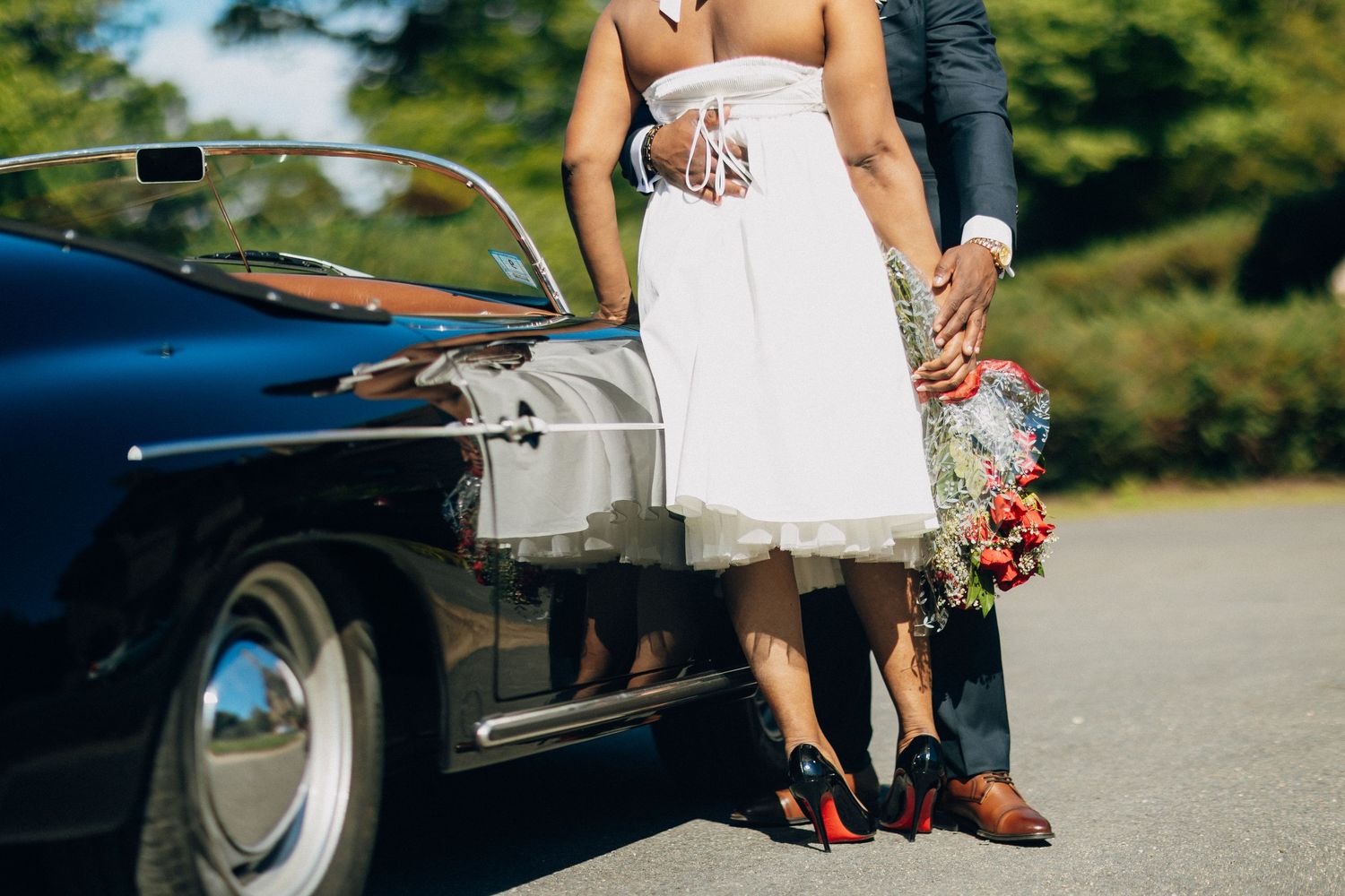 Close-up of Black bride in white dress and groom holding a colorful bouquet beside a vintage black car.