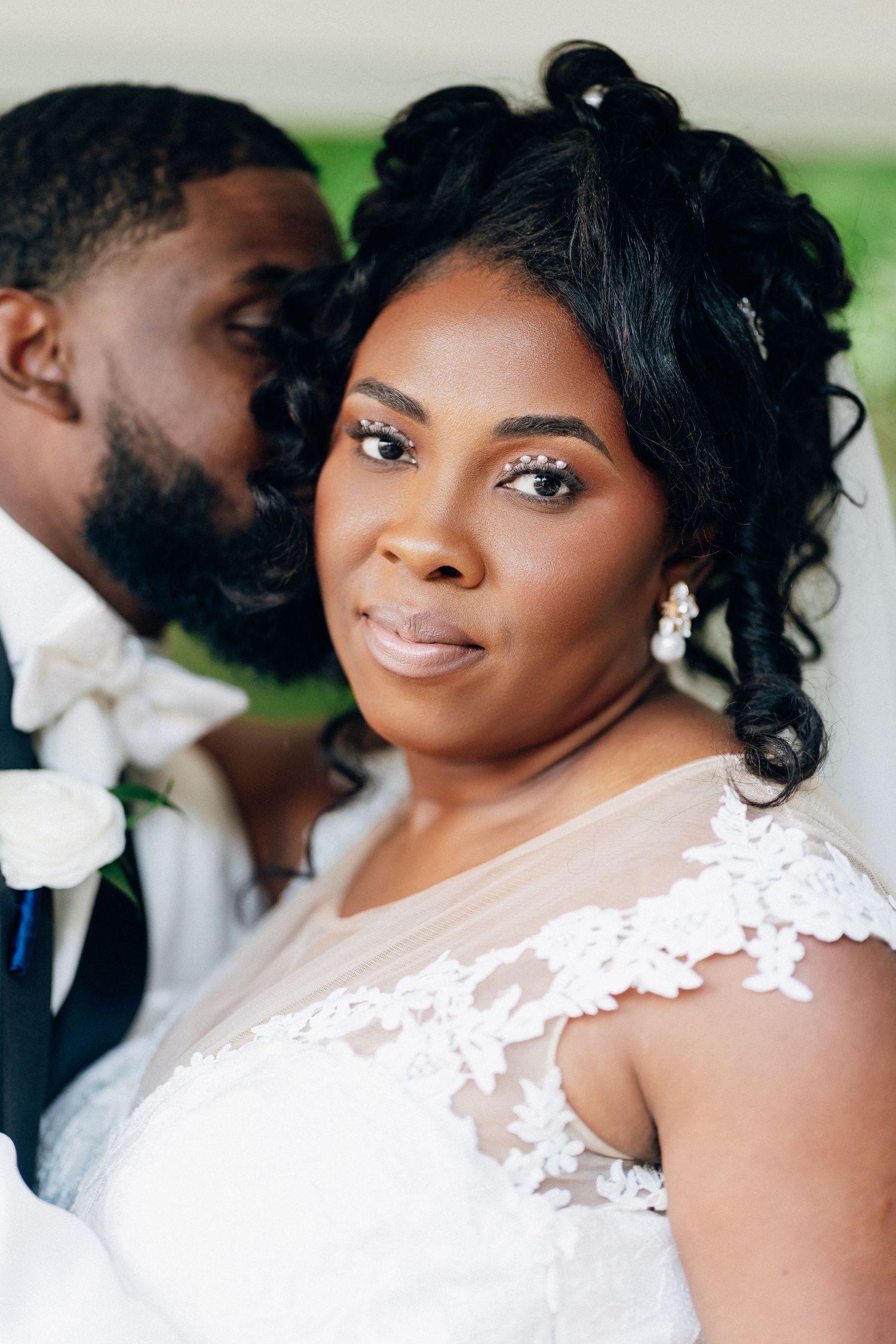 A bride in lace wedding dress smiles at camera as groom in navy suit kisses her cheek at wedding ceremony.