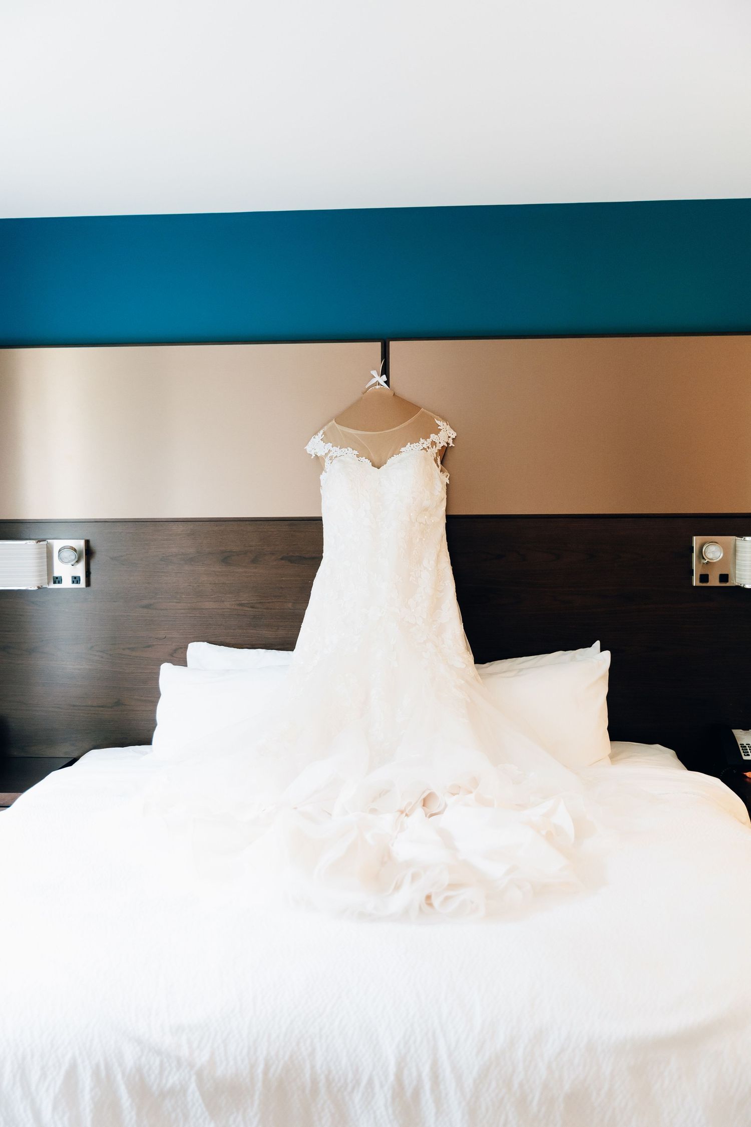 Elegant white lace wedding dress hanging on hanger above hotel bed with white pillows.