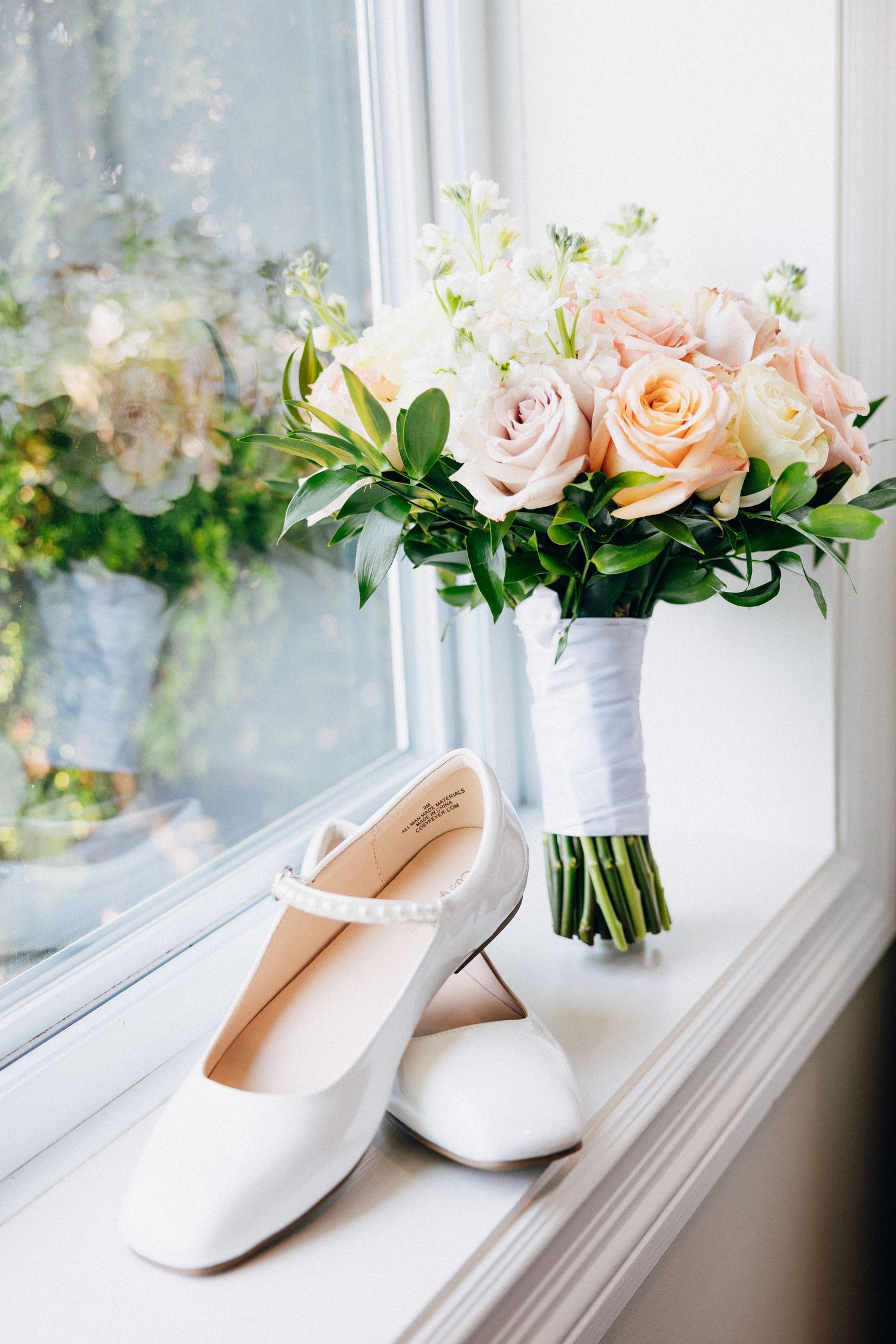 White bridal shoes and a blush pink rose bouquet with greenery displayed on a sunlit window sill.