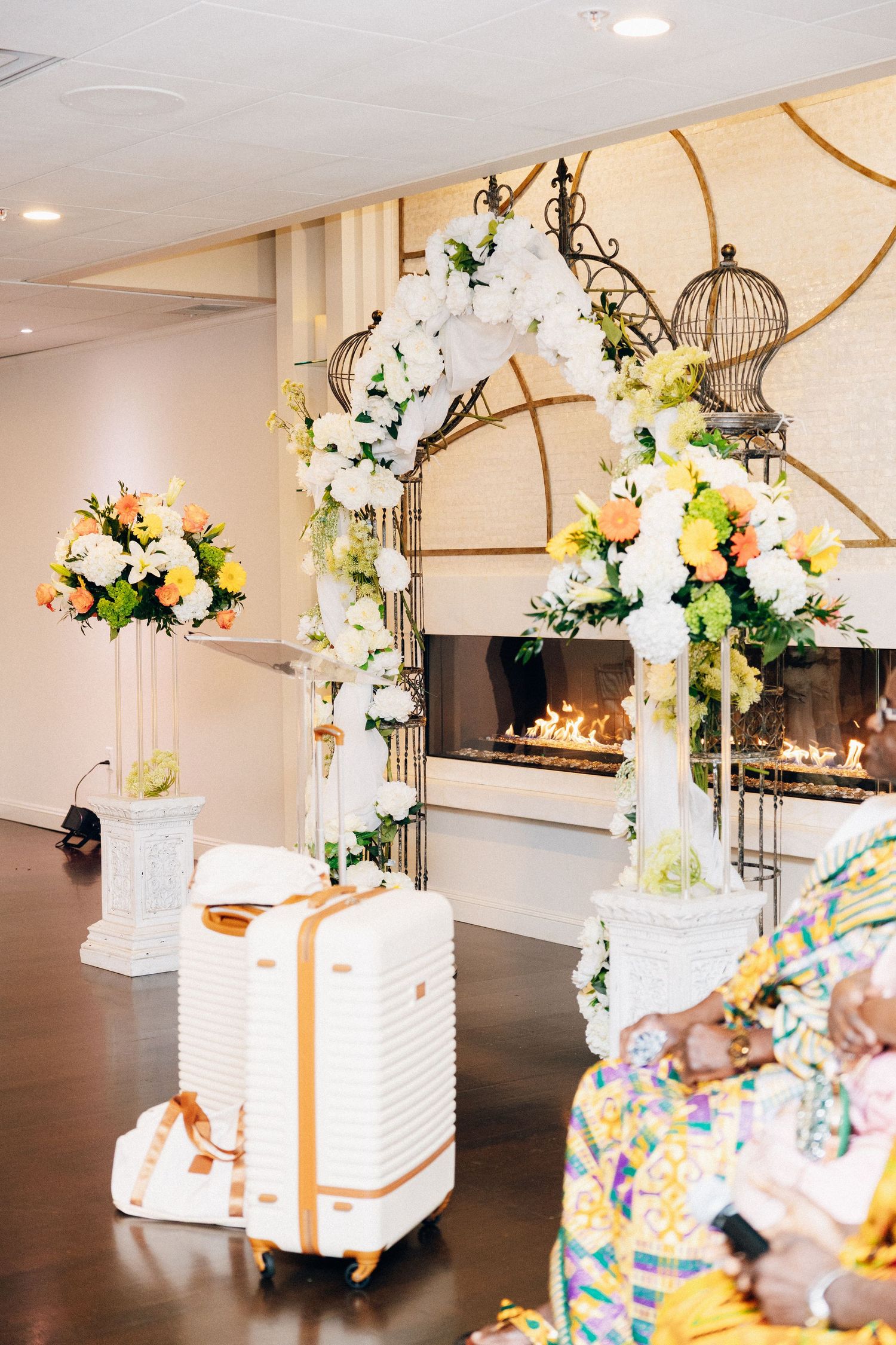 Elegant wedding arch adorned with white and yellow flowers beside a fireplace with birdcages and a white luggage.