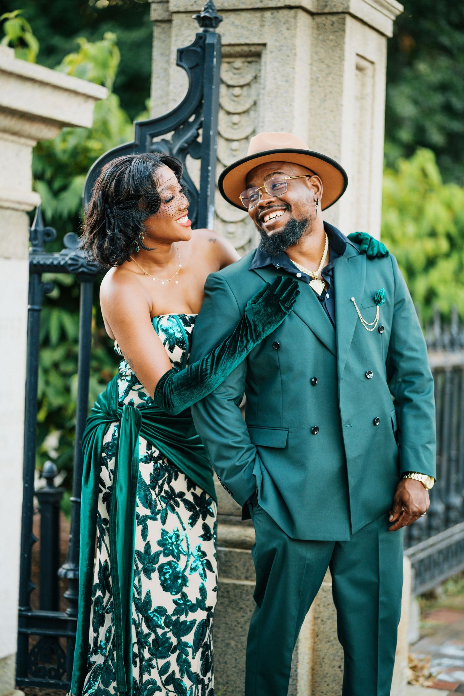 Smiling Black couple in coordinated emerald green outfits near ornate iron gate.