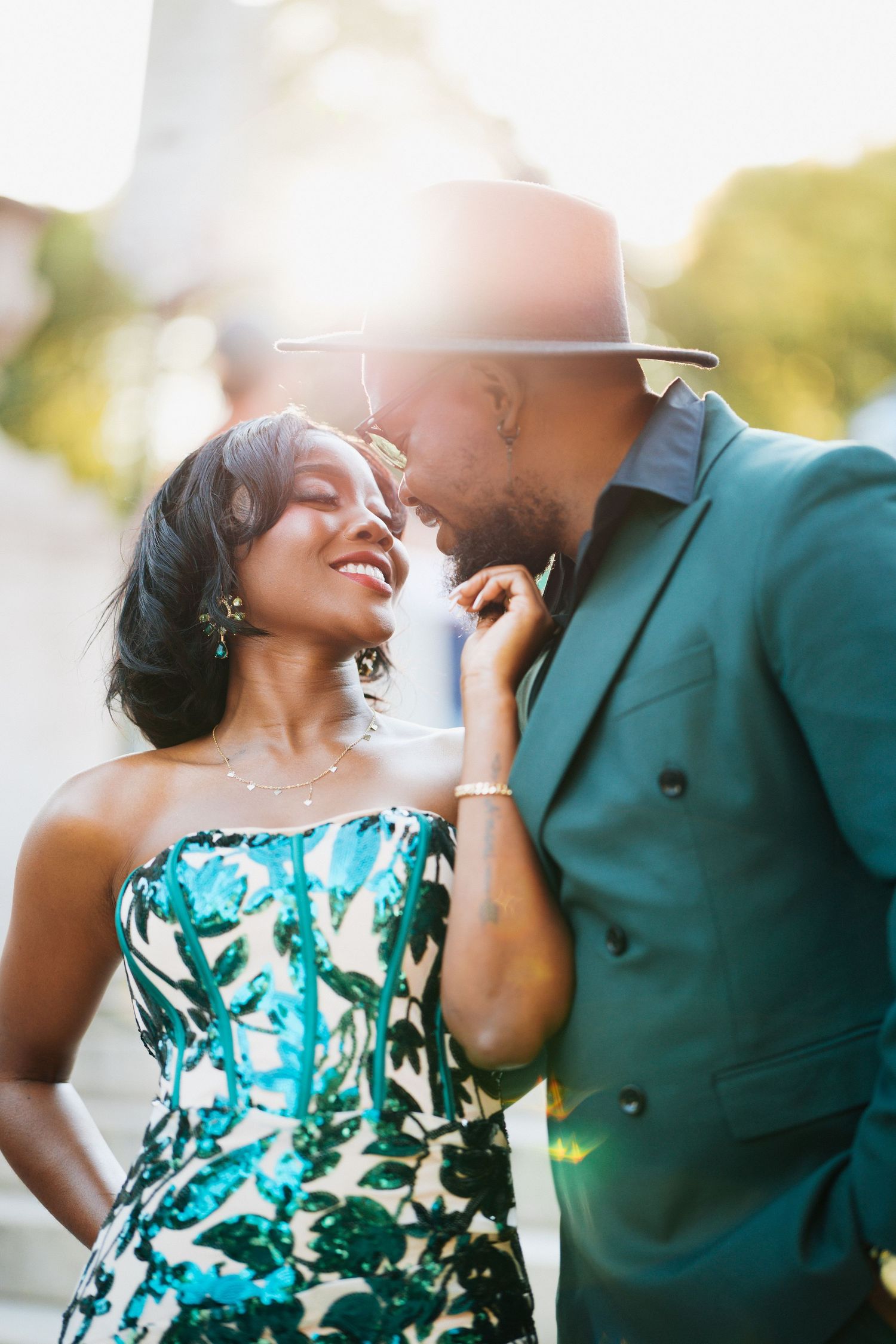 A stylish couple in teal outfits share a romantic moment outdoors, with the woman smiling in a sequined dress.