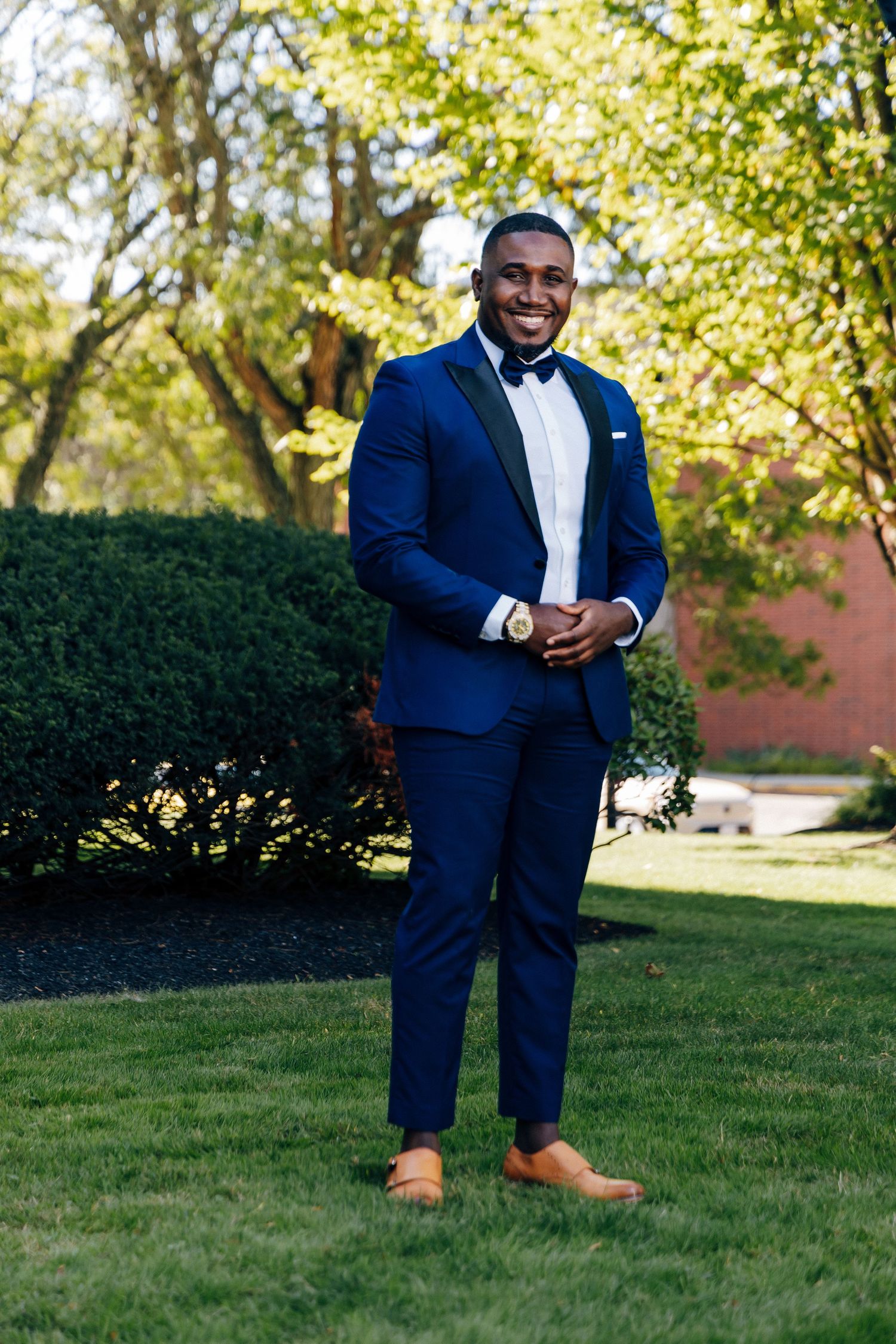 Smiling groom in navy blue tuxedo with black bow tie and tan loafers posing in lush garden.