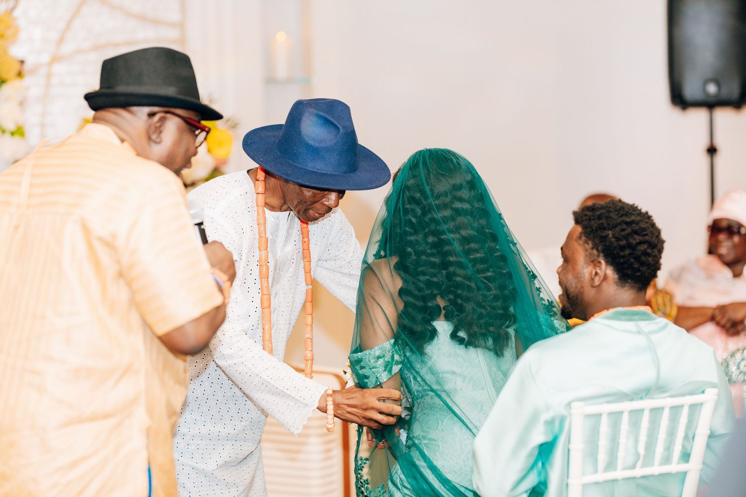Guests in traditional African attire gather around a bride in teal veil during a cultural wedding ceremony.