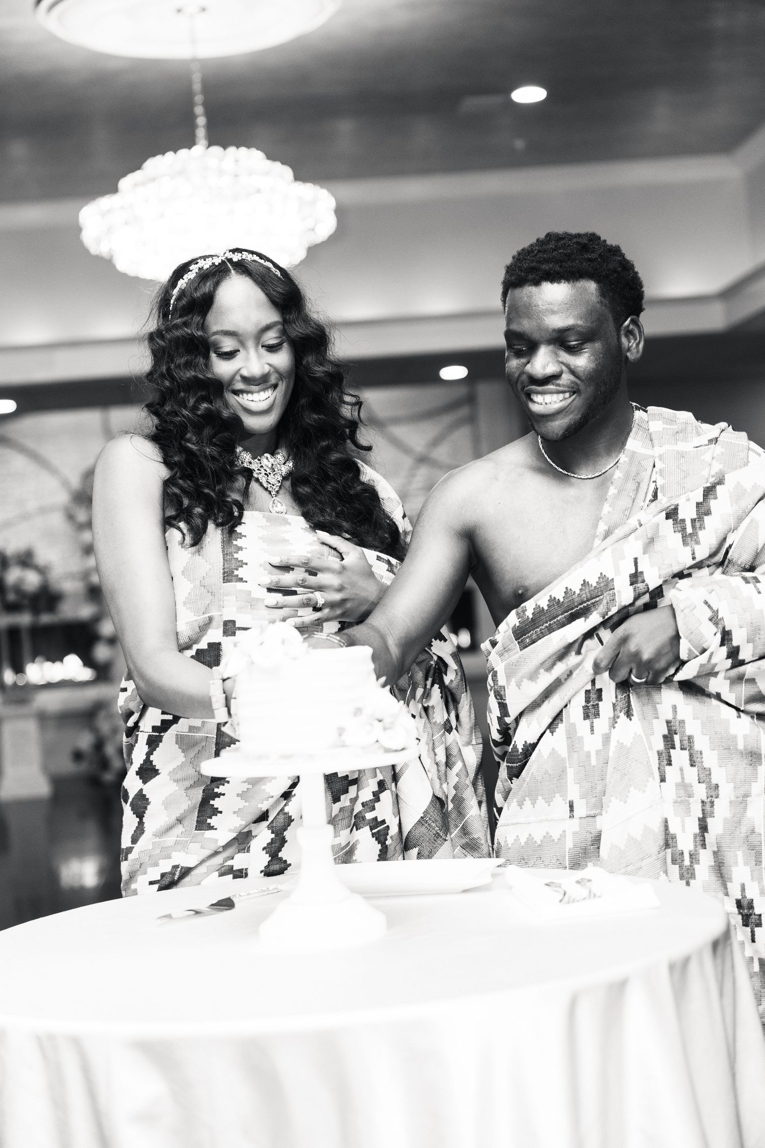 Smiling couple in Kente attire cutting wedding cake at elegant reception.