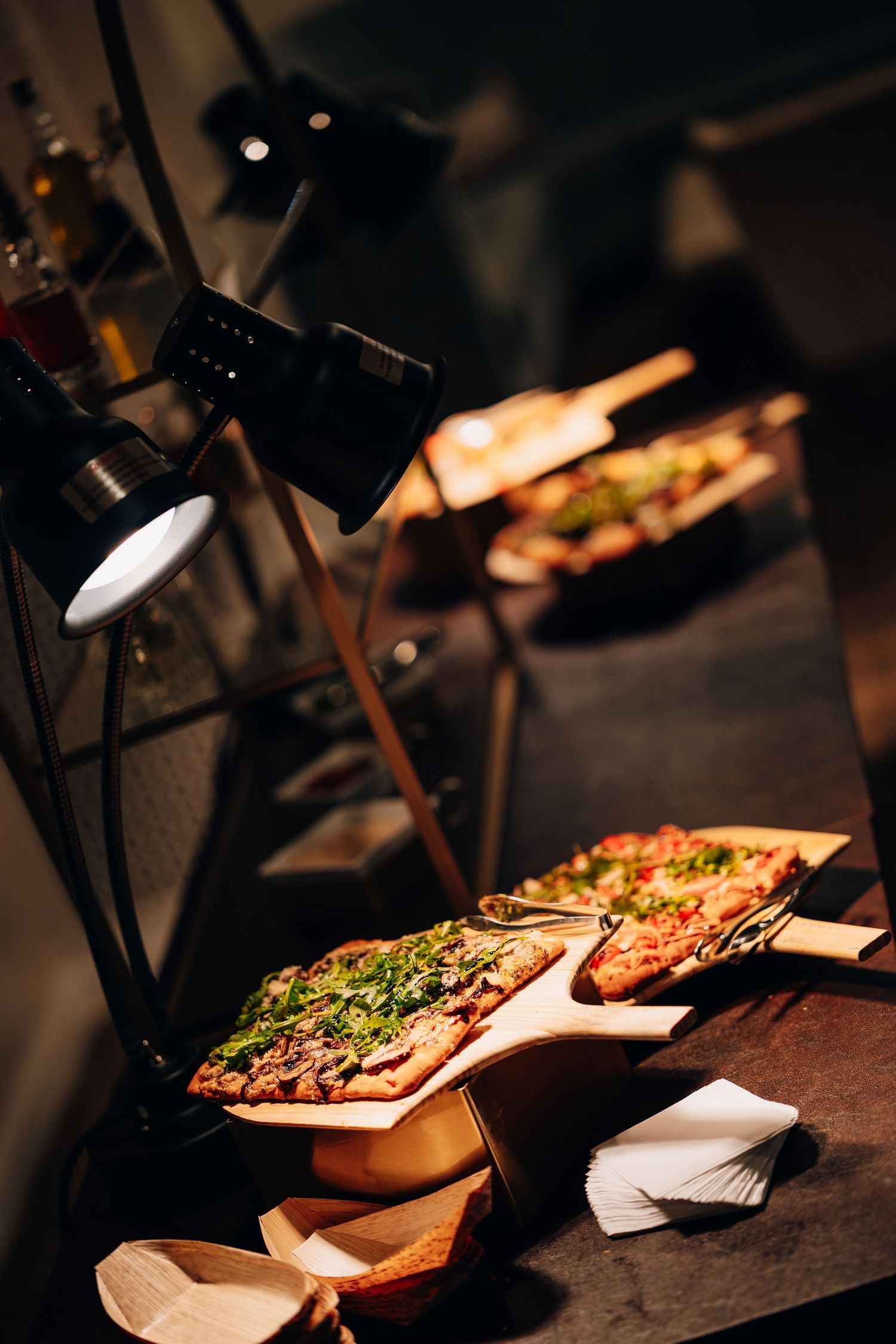 Pizza slices on wooden boards displayed under heat lamps at catering station.