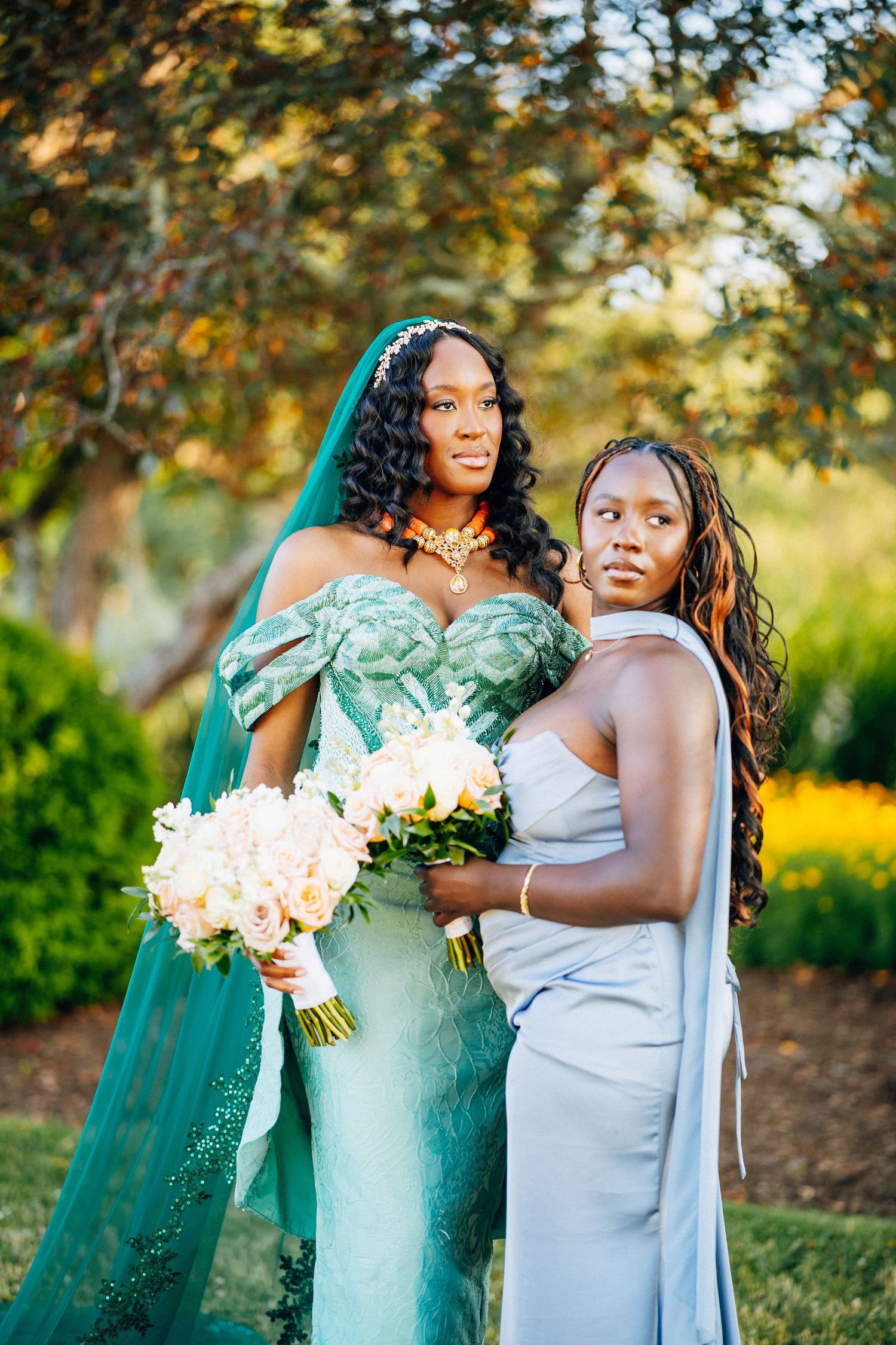 Nigerian bride in teal beaded gown with coral jewelry poses with bridesmaid in silver dress outdoors.