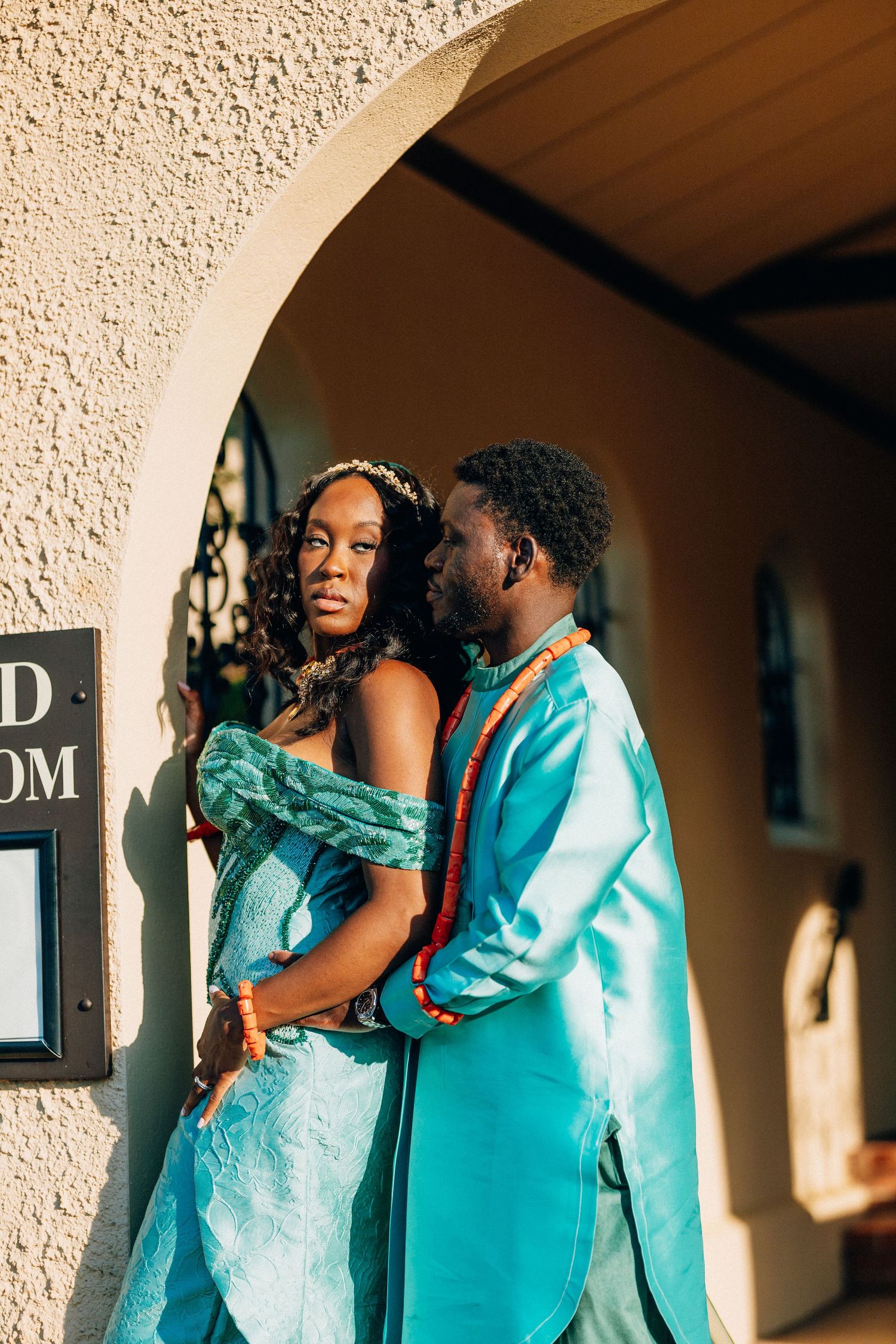 A Black couple in matching teal traditional attire pose romantically under an arched doorway.