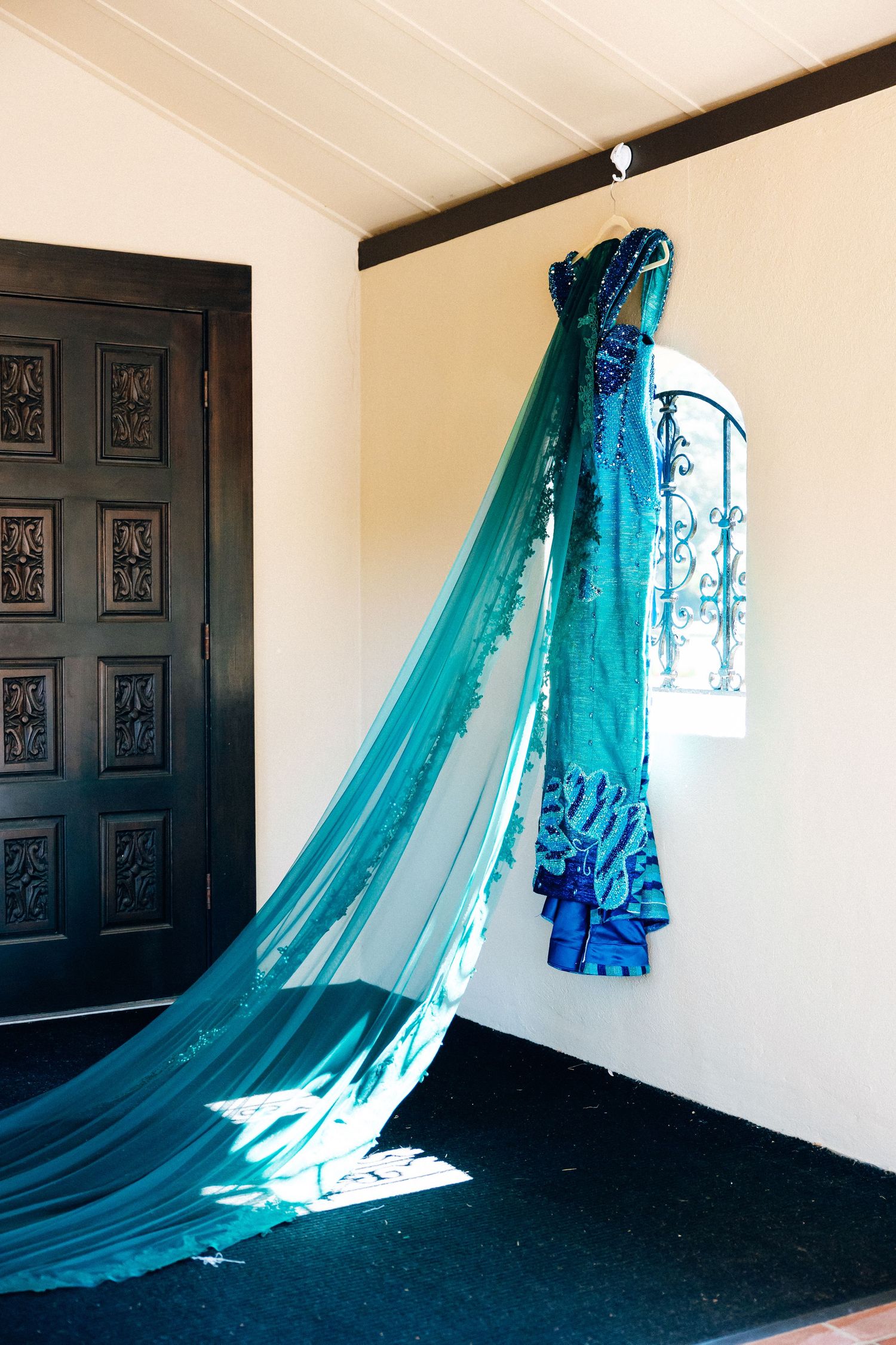 Teal strapless gown with lace train hanging on wall near ornate dark door and arched window.