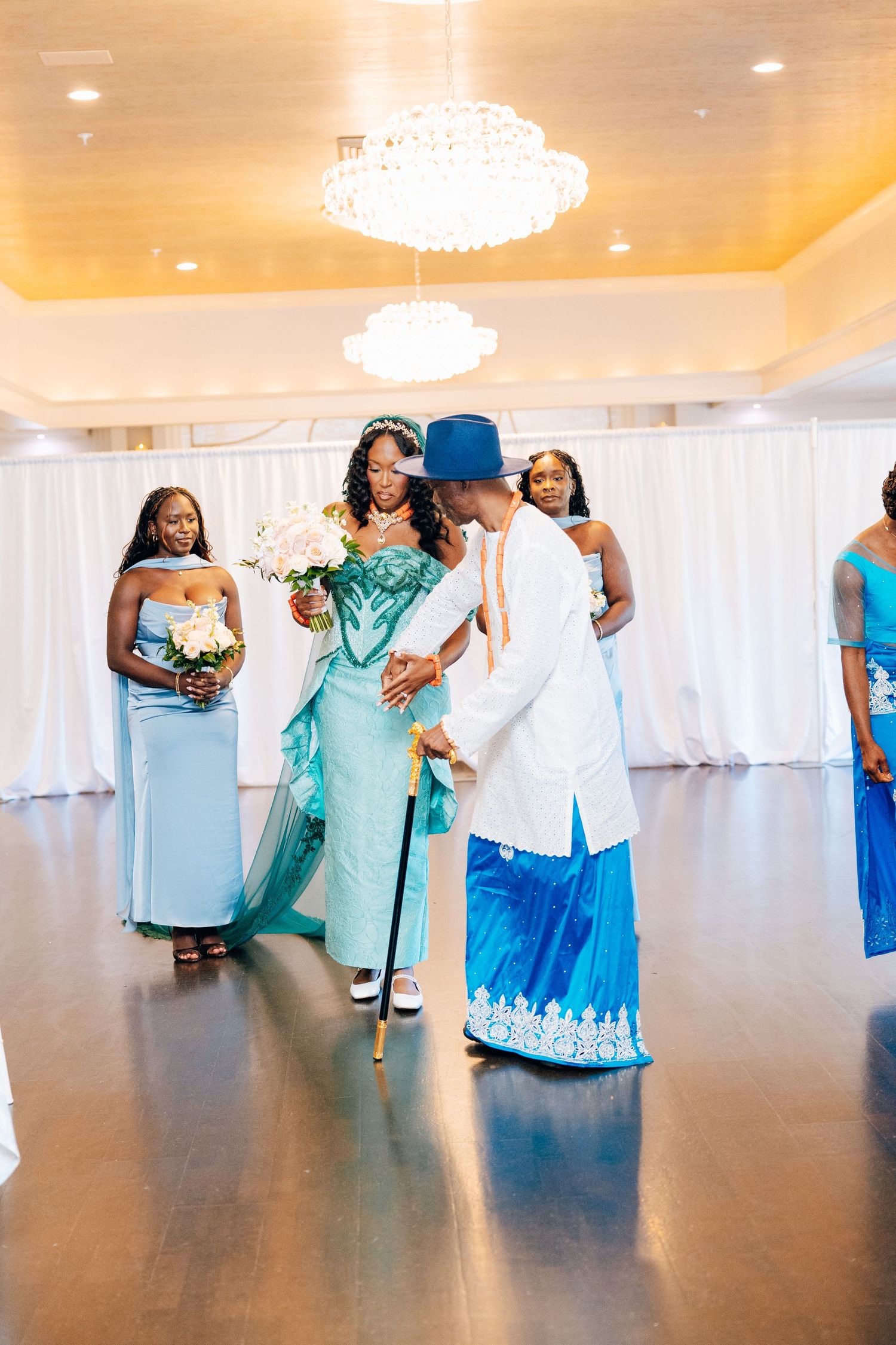 Bride in teal African dress walks with elder man in blue traditional attire and cane during wedding processional.