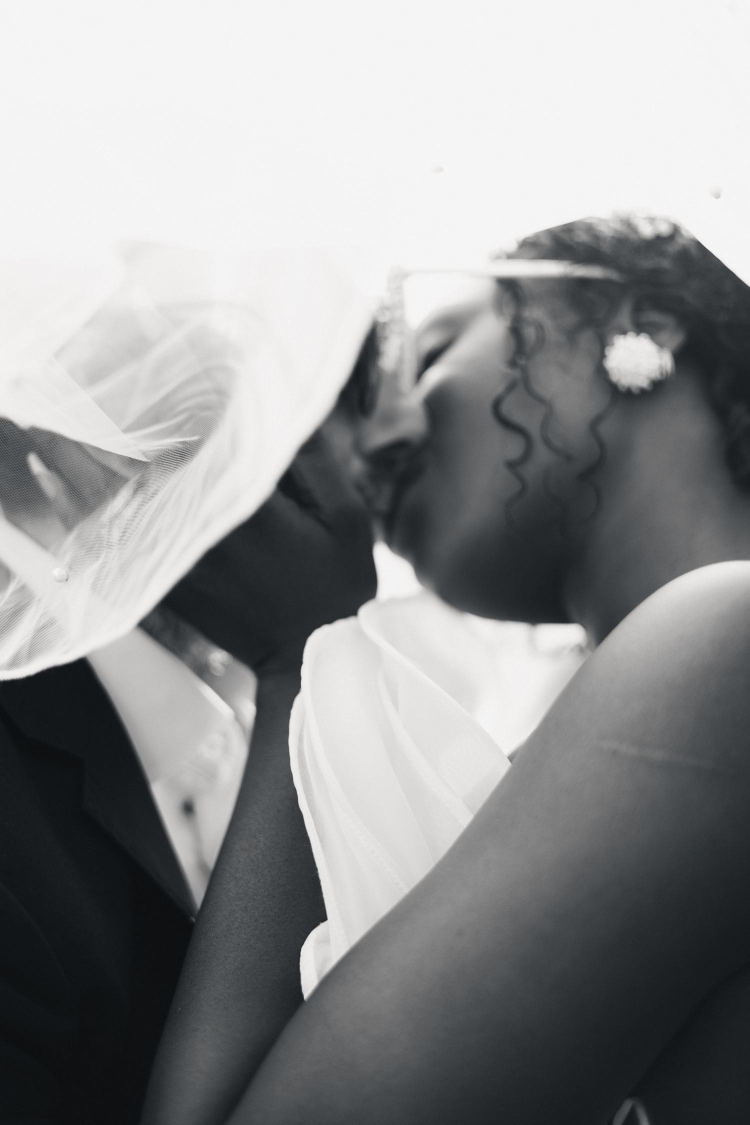 A black and white close-up of an interracial bride and groom sharing a tender kiss under a wedding veil.