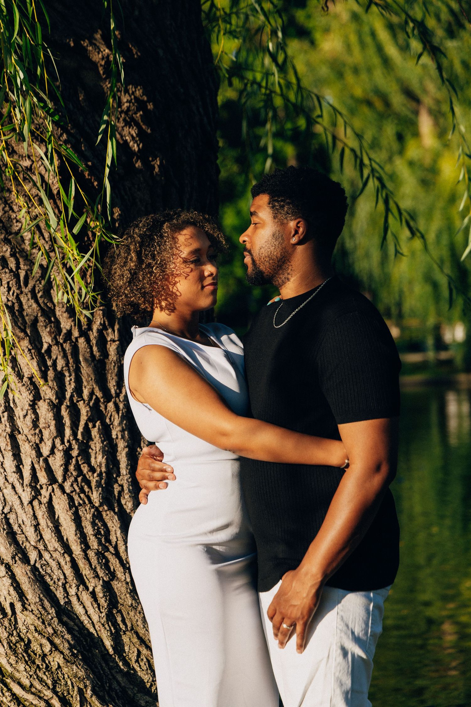 A couple embraces tenderly near a large tree by a serene pond, the woman in white and man in black shirt.