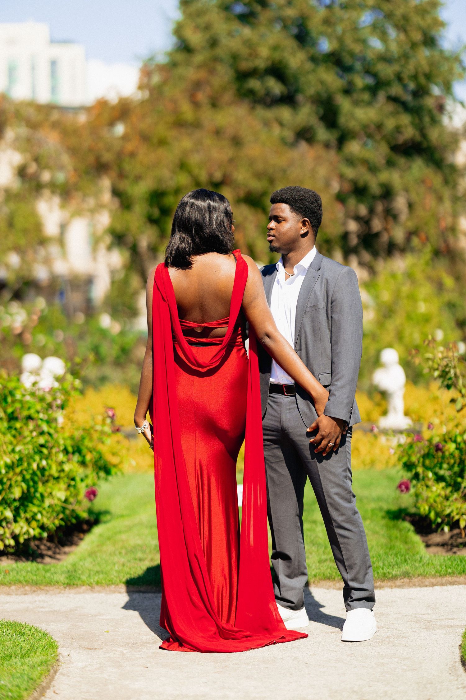 Elegant couple in a garden, woman in red gown with cape, man in gray suit, sharing a romantic moment.