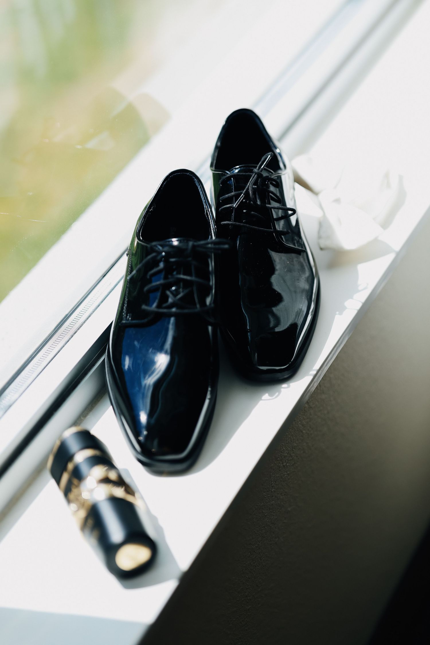 Shiny black patent leather dress shoes with cologne bottle displayed on white window sill.