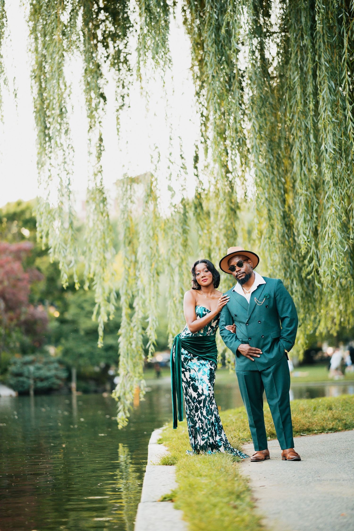Sophisticated Black couple in emerald green attire posing under weeping willow by pond.
