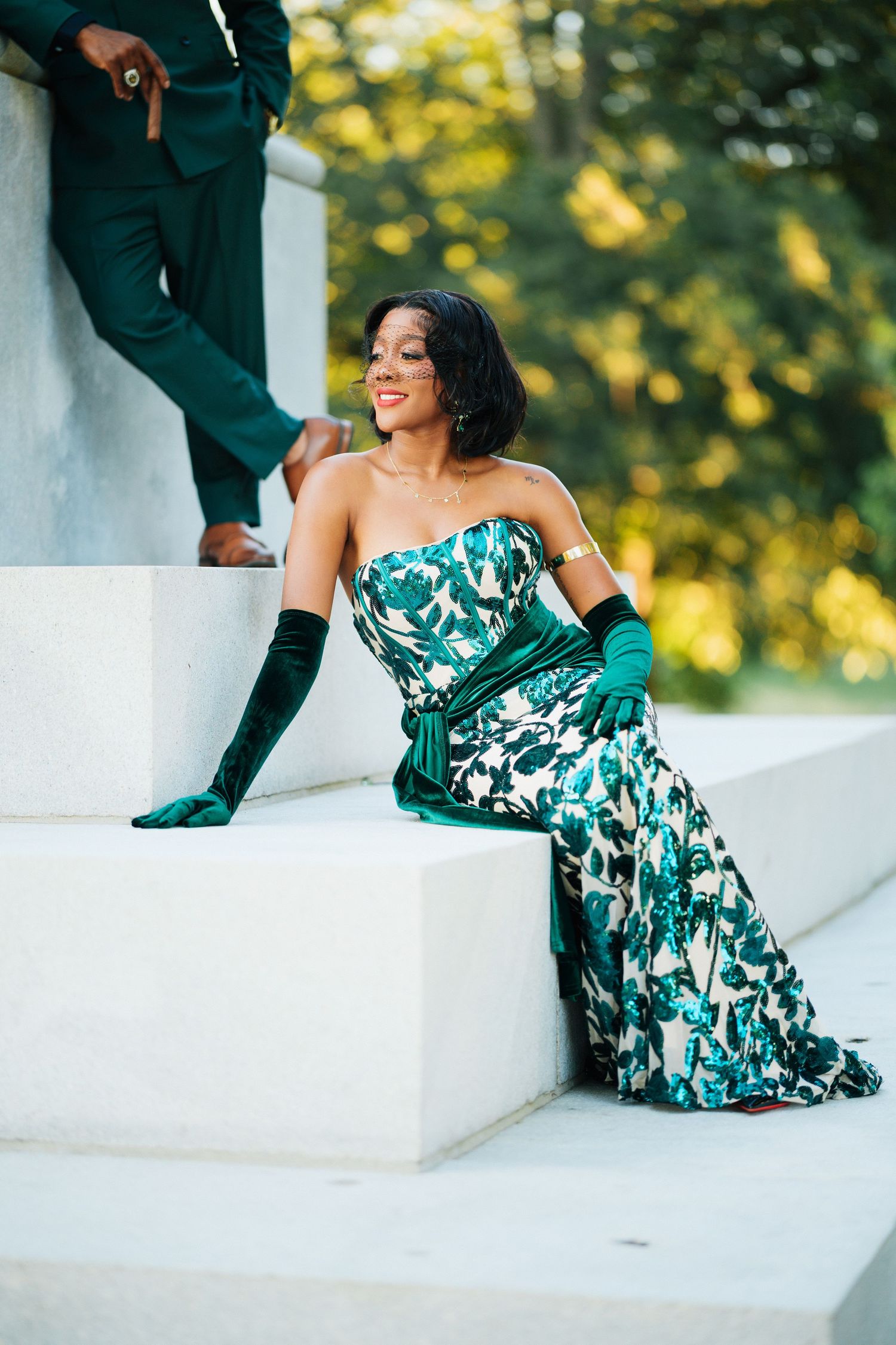 Glamorous Black woman in green floral gown with velvet gloves posing on white marble steps outdoors.