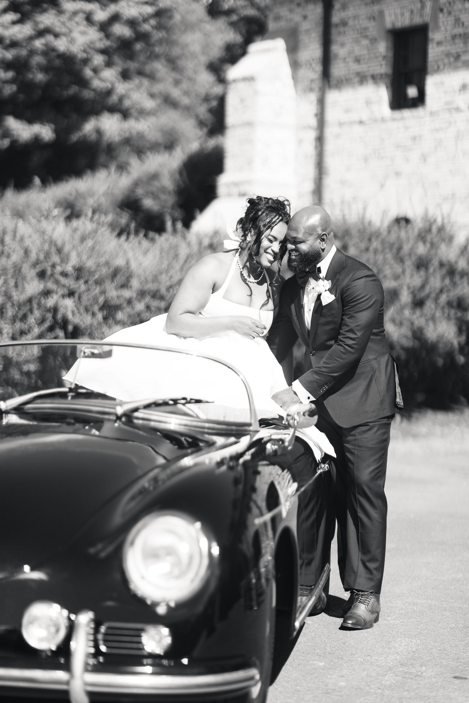 Bride and groom share intimate moment leaning against vintage classic car in black and white wedding photo.