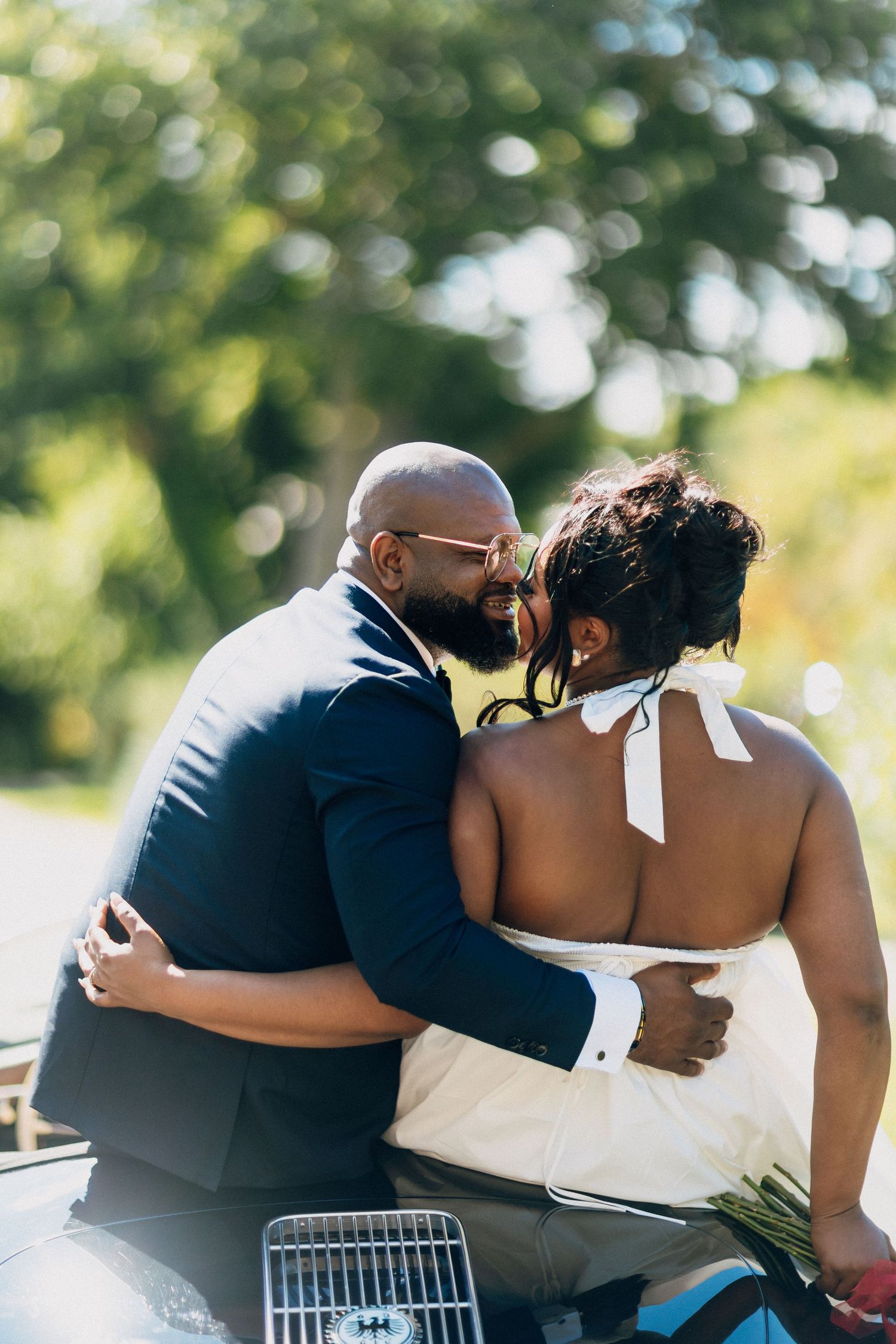 Groom in navy suit embraces his bride from behind while seated in a vintage convertible on wedding day.