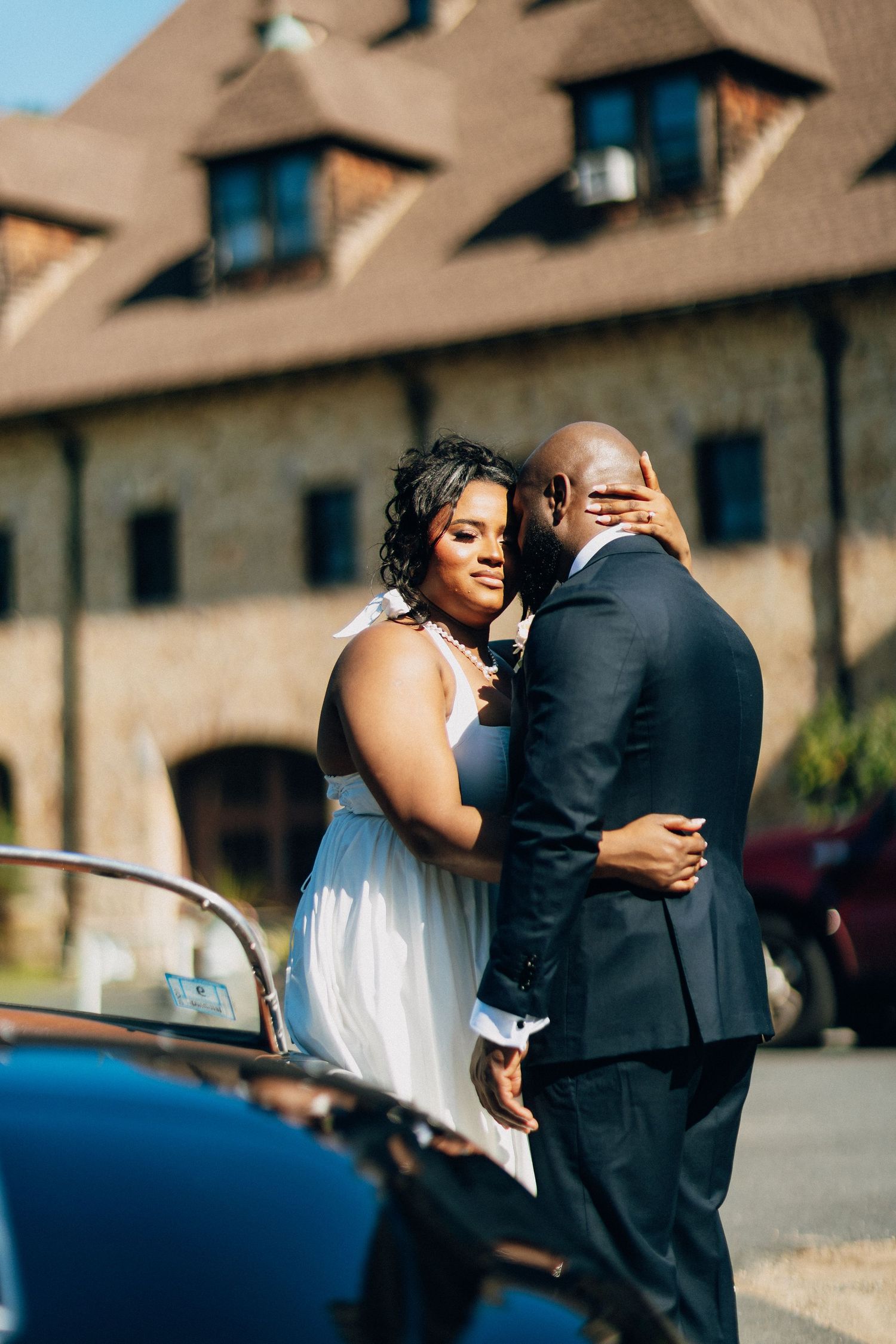 A bride and groom share a tender embrace outside a rustic wedding venue beside a vintage car.