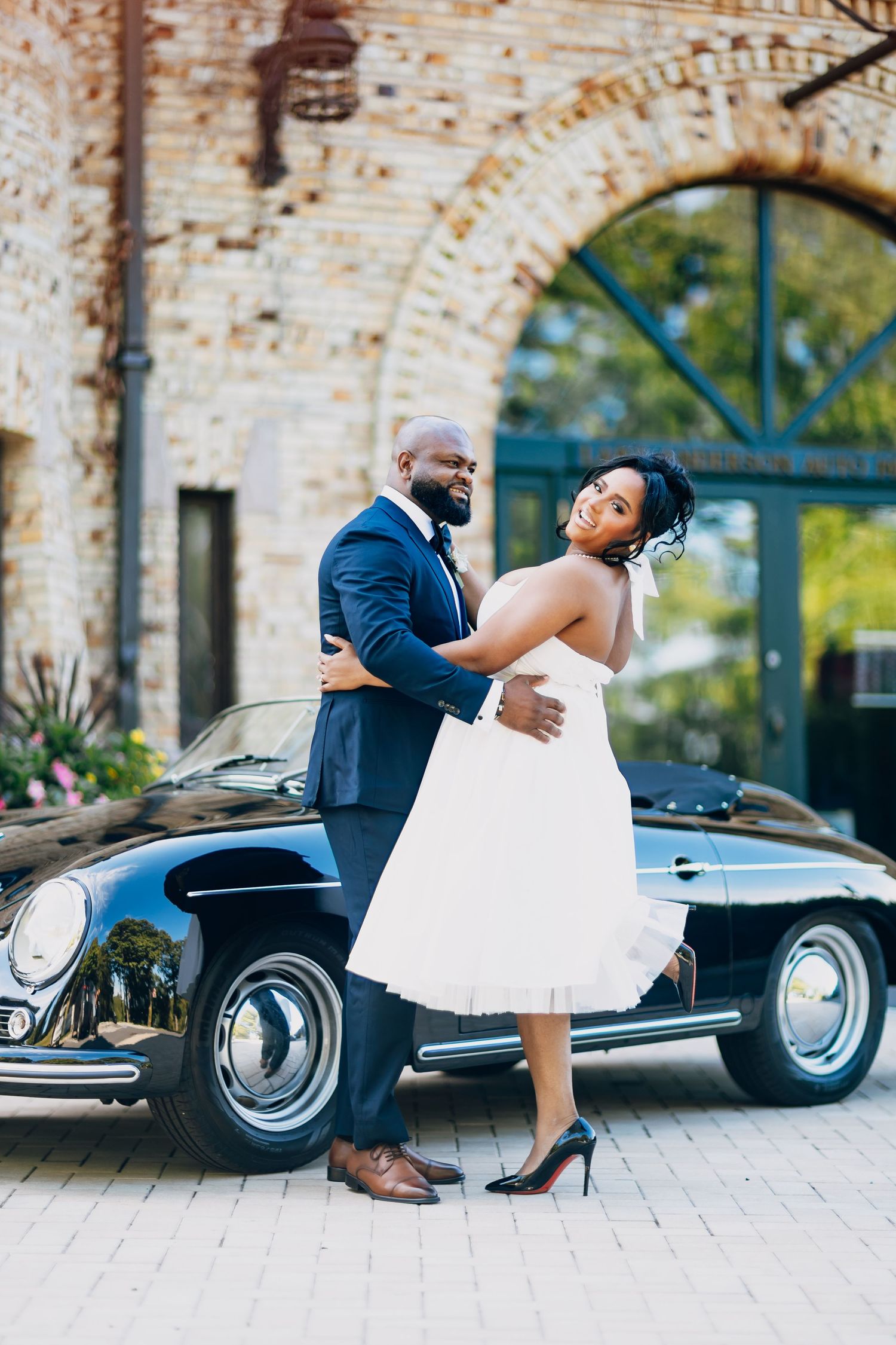 Bride and groom embrace beside a classic black Porsche convertible outside a brick arch venue.