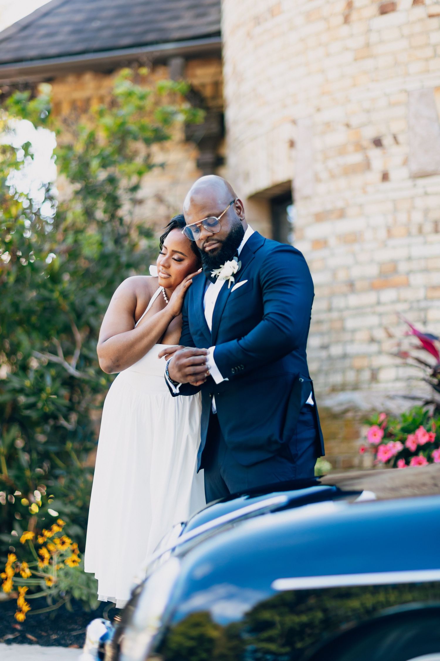 A bride leans tenderly against her groom near a classic black car at a stone venue.