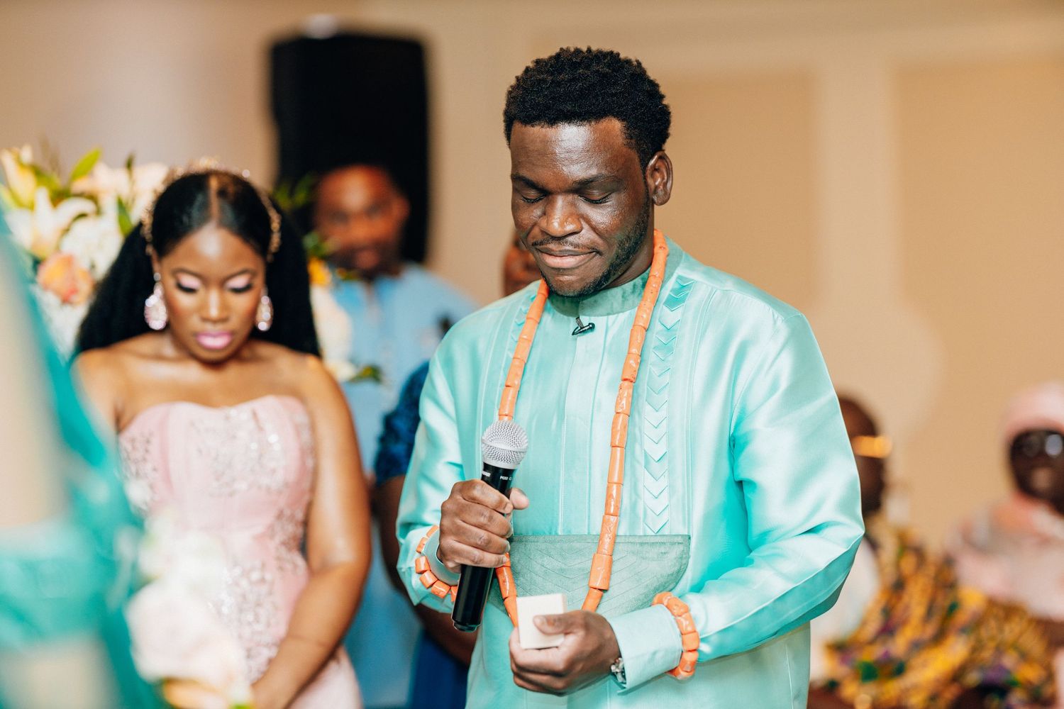 Man in teal African attire with orange beads holds microphone at elegant wedding reception ceremony.