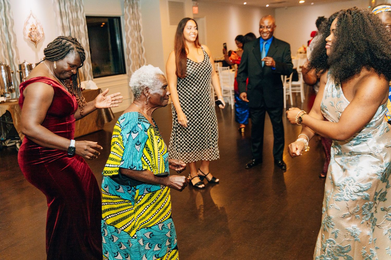 Guests dancing joyfully at an indoor celebration, featuring women in colorful attire on a wooden dance floor.