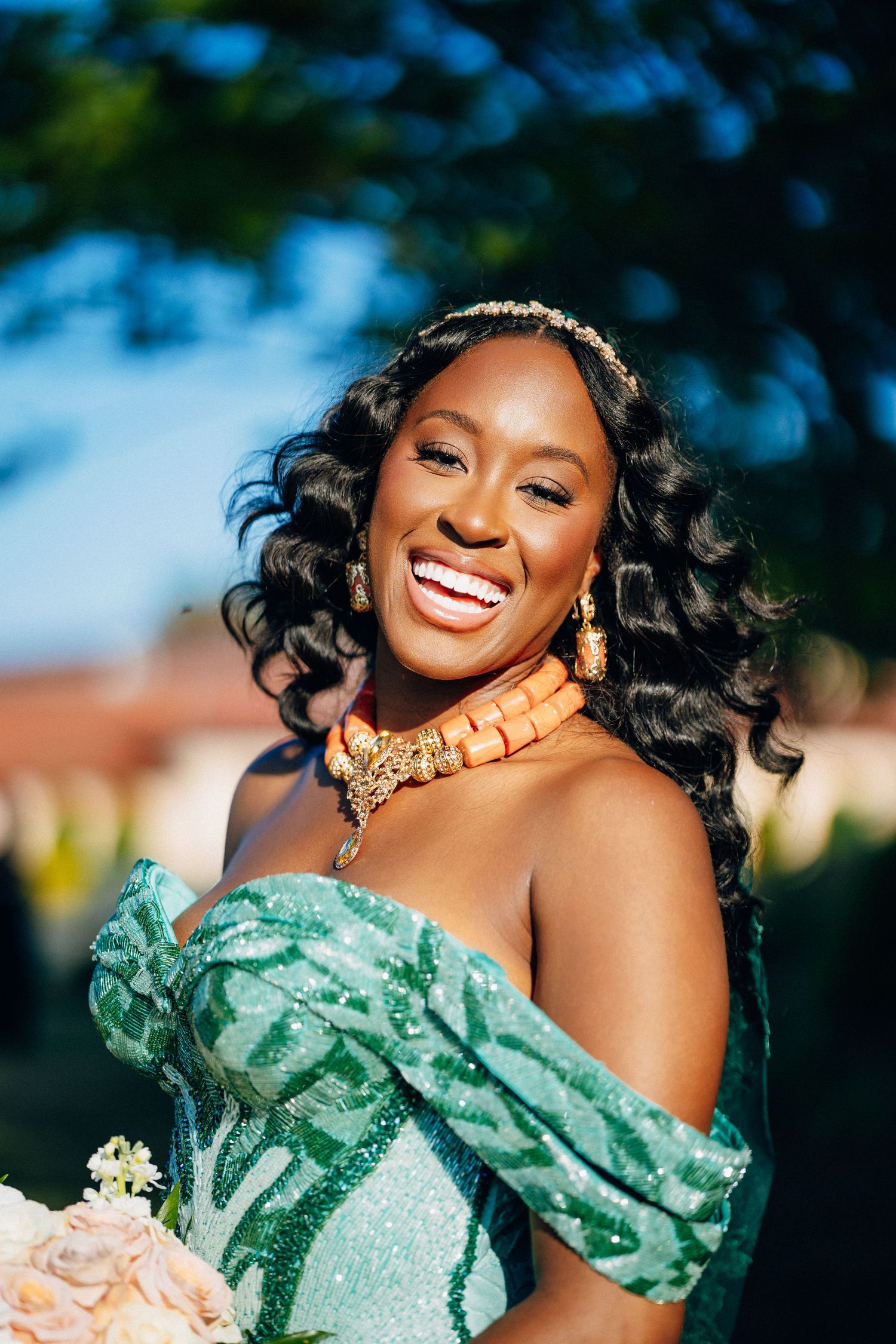 Smiling bride in sparkling green off-shoulder gown with orange coral jewelry and headpiece holding bouquet outdoors.