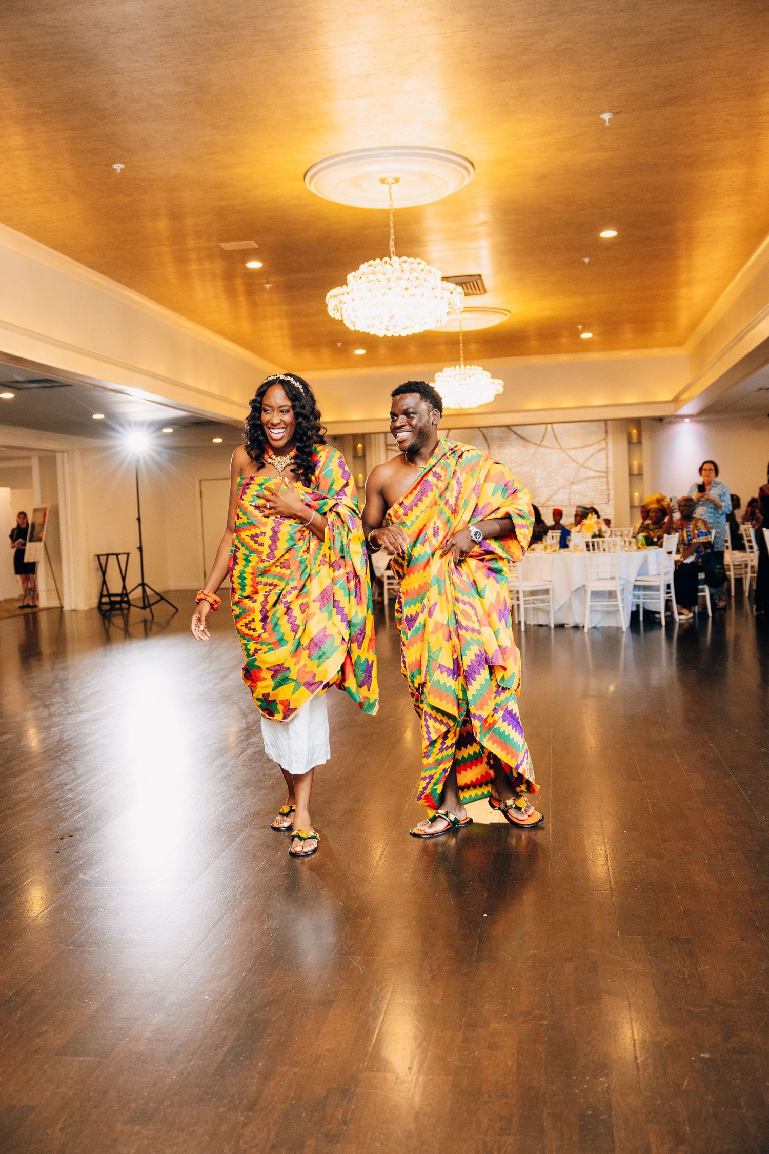 Couple dancing in colorful African kente cloth outfits on a wooden dance floor at a wedding reception.
