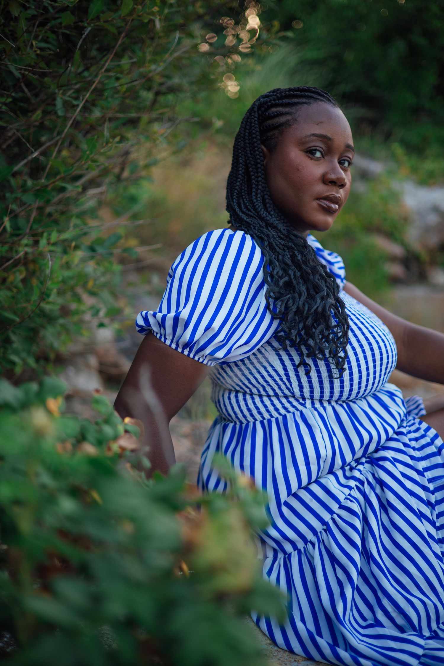 A Black woman in a blue and white striped dress with braided hair poses outdoors among lush green foliage.