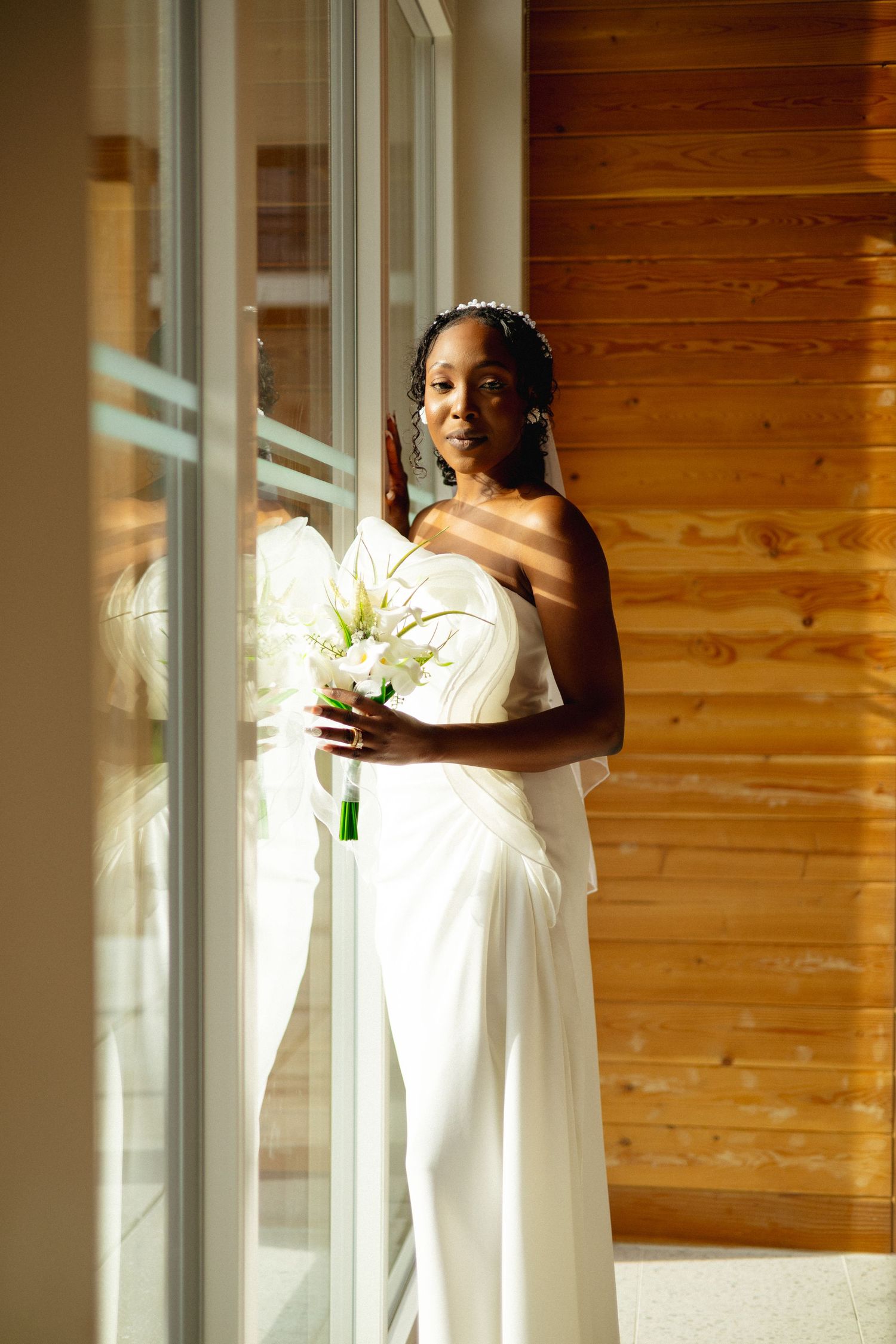 Elegant Black bride in white strapless gown holding calla lily bouquet, standing by sunlit window with wooden wall backdrop.