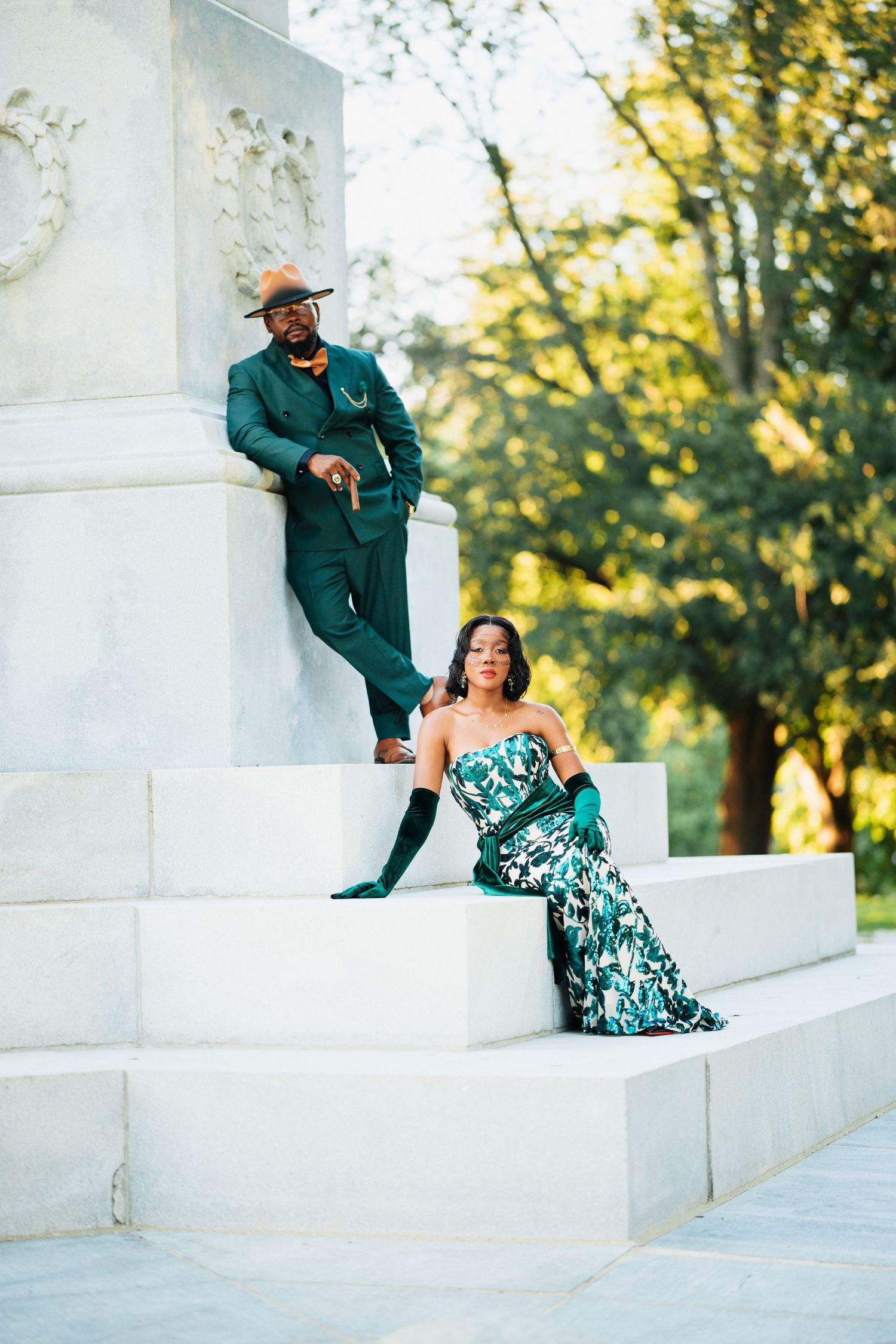 Elegant couple in matching green outfits posing on white marble steps near a monument surrounded by lush trees.