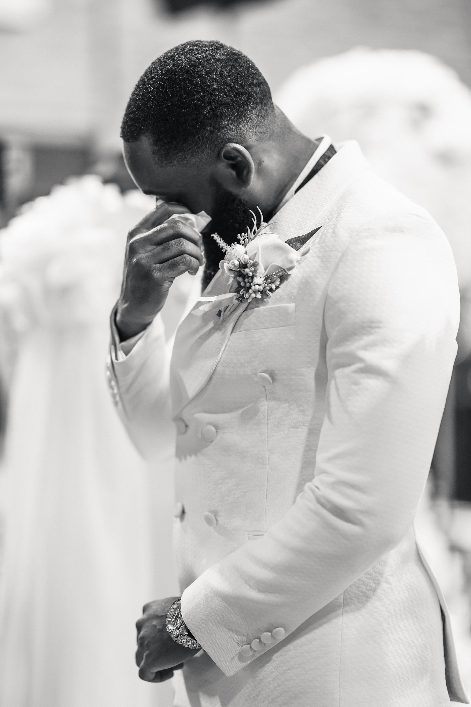 Emotional groom in white tuxedo wiping tears during wedding ceremony, wearing boutonniere, black and white photo.