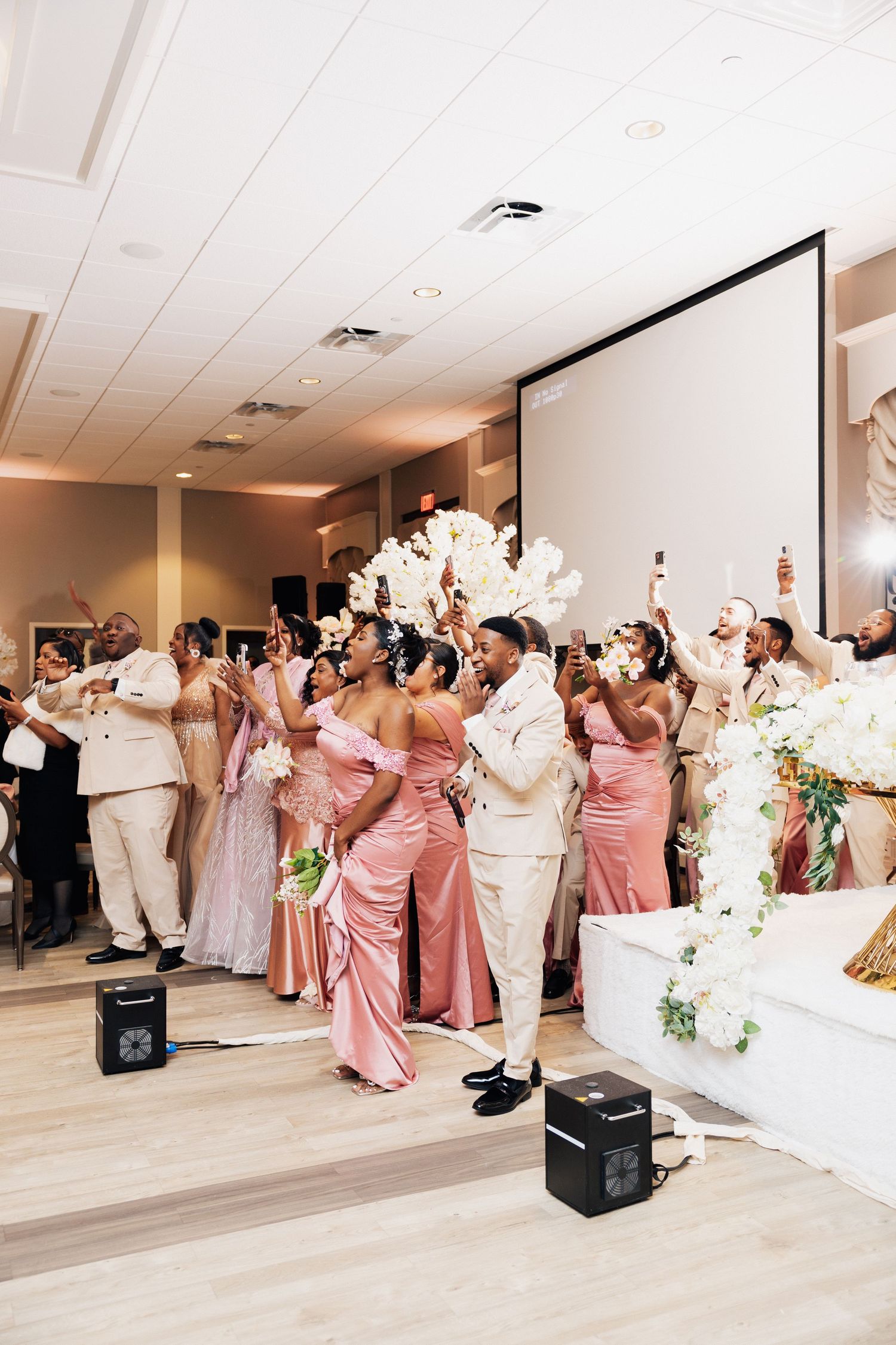 Wedding party in pink gowns and ivory suits celebrate joyfully at an elegant indoor reception venue.