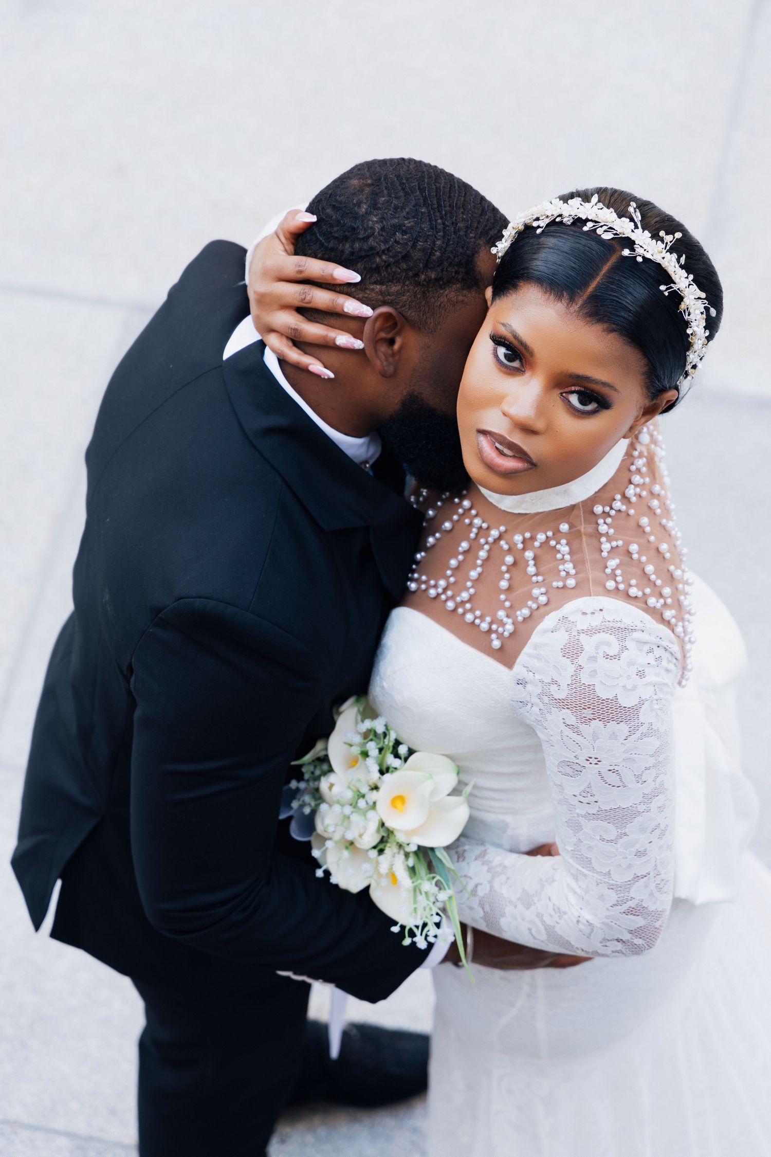 Elegant Black bride in pearl-adorned white lace gown embraces groom in black suit, holding white floral bouquet outdoors.