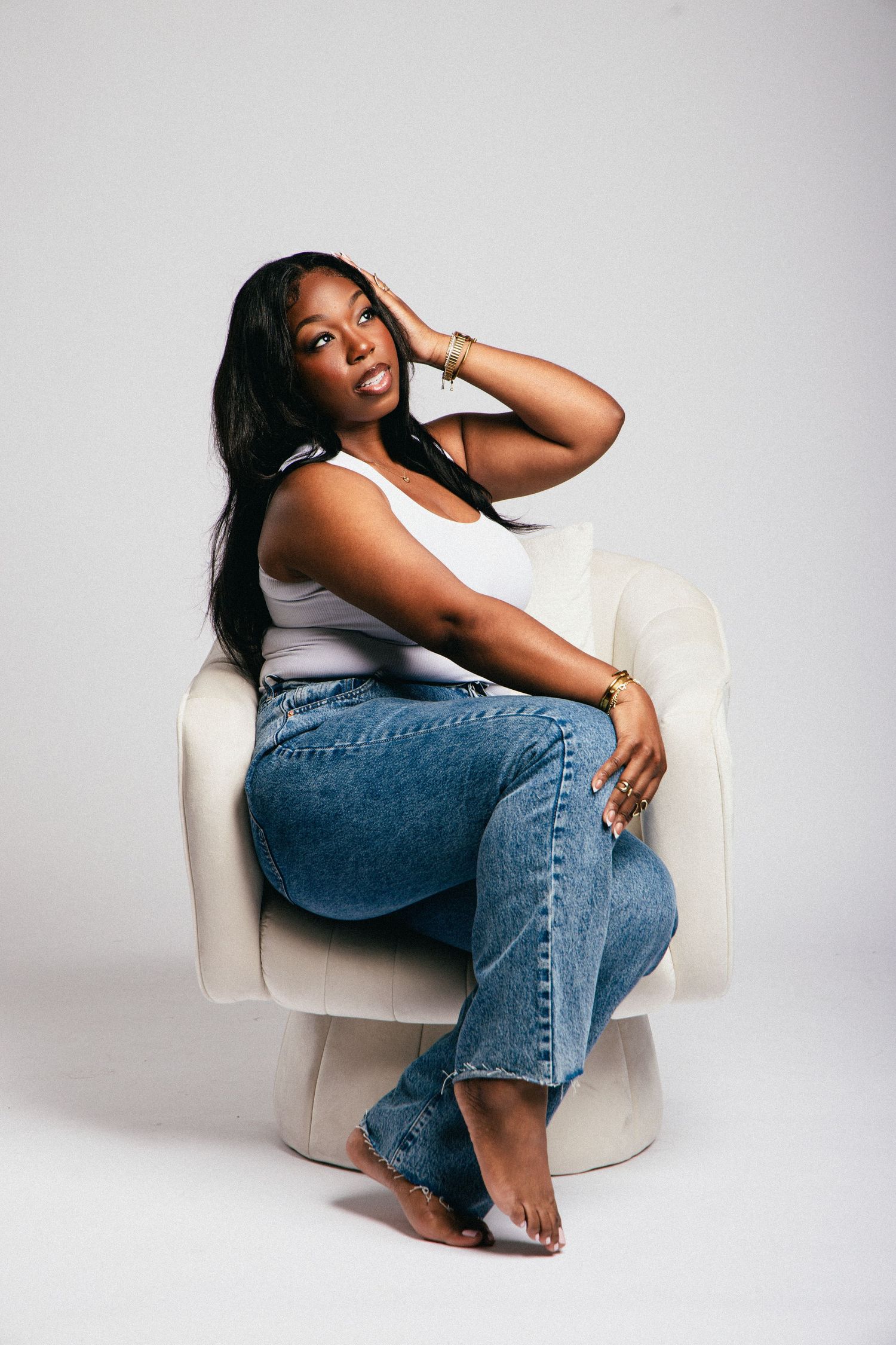 A woman in a white tank top and blue jeans sits relaxed in a cream chair, posing confidently barefoot.