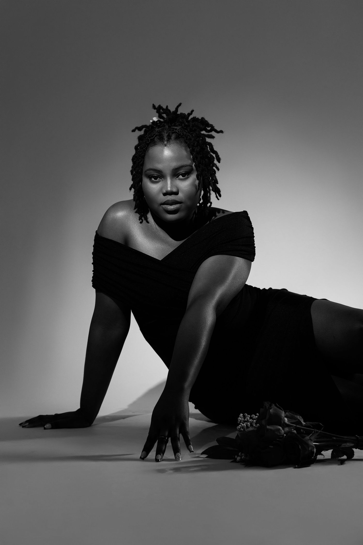 Black and white studio portrait of a woman in black off-shoulder top posing gracefully on the floor.